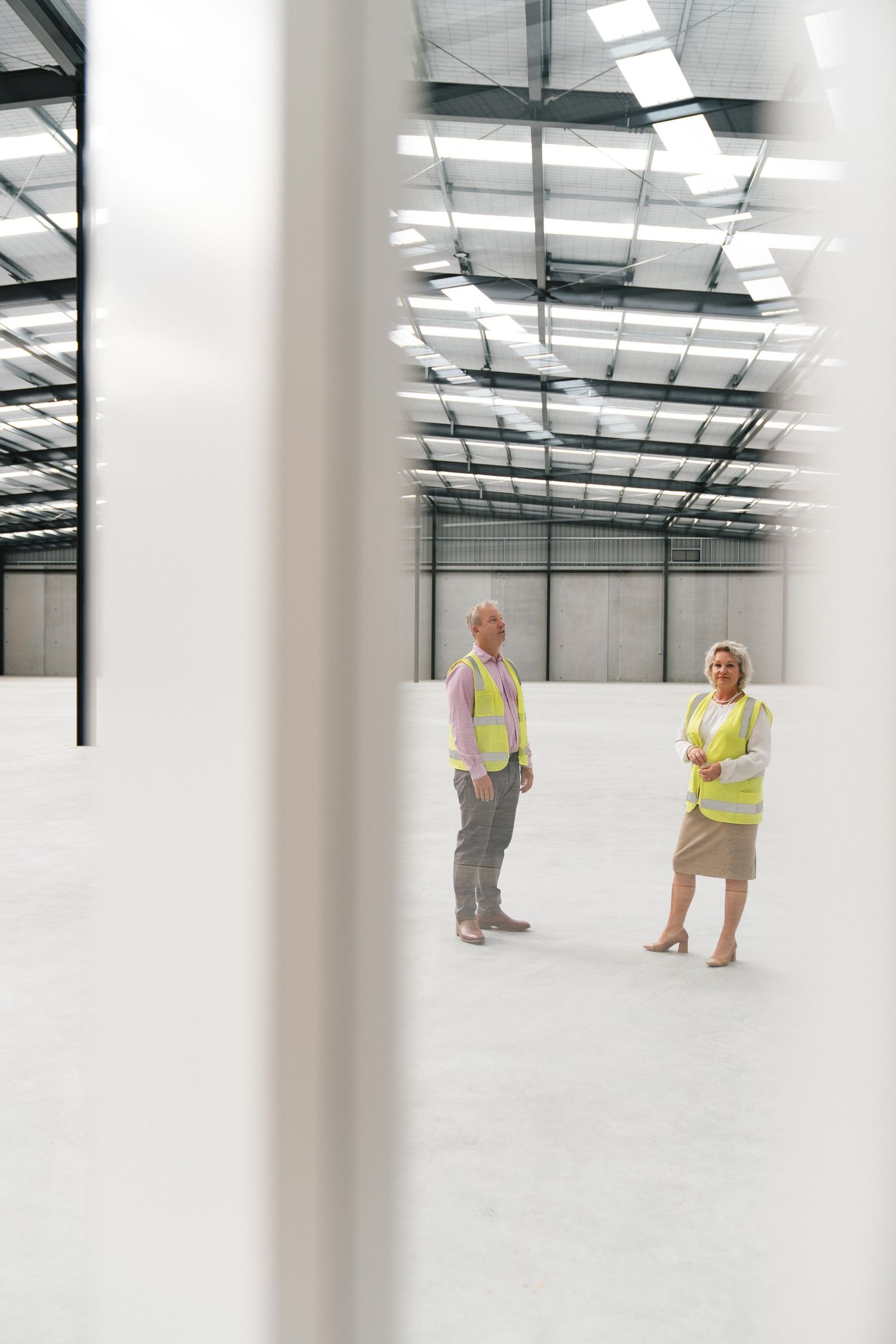 Two workers in safety vests observe an empty industrial warehouse space with skylights and exposed ceiling structure.