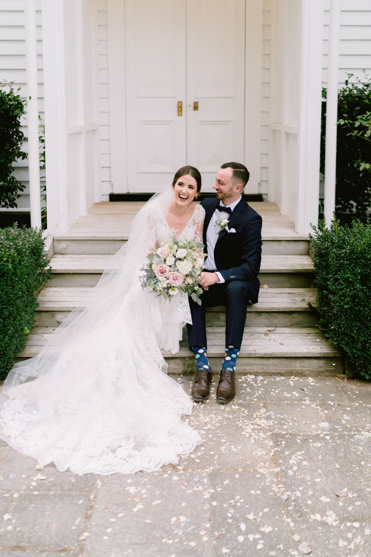 A couple sits on white steps scattered with flower petals in front of a classic white columned entrance.