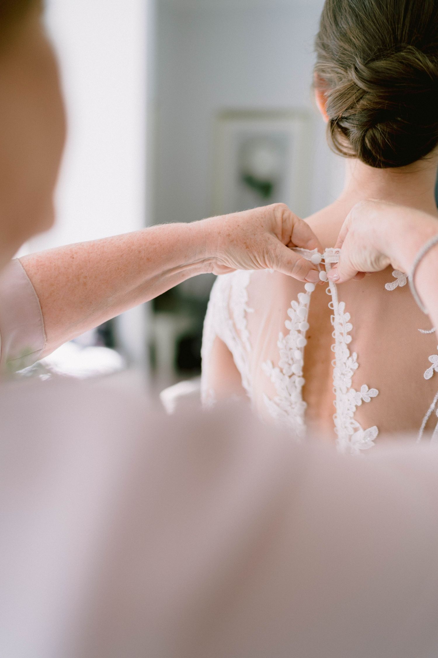Someone helps button up the delicate lace back of an elegant white wedding dress with intricate floral details.