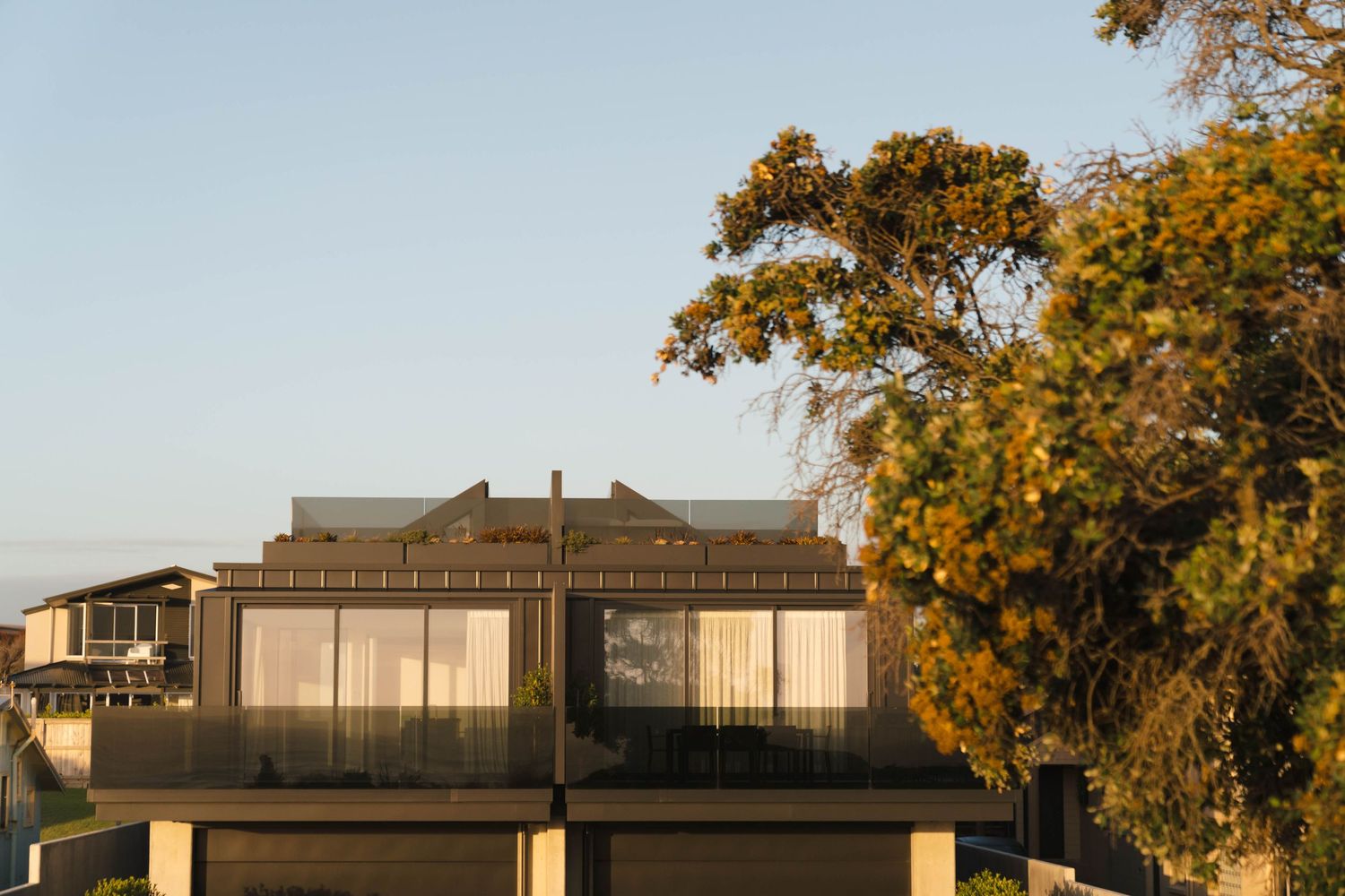 Modern multi-level house with large windows set against autumn foliage and blue sky at sunset.