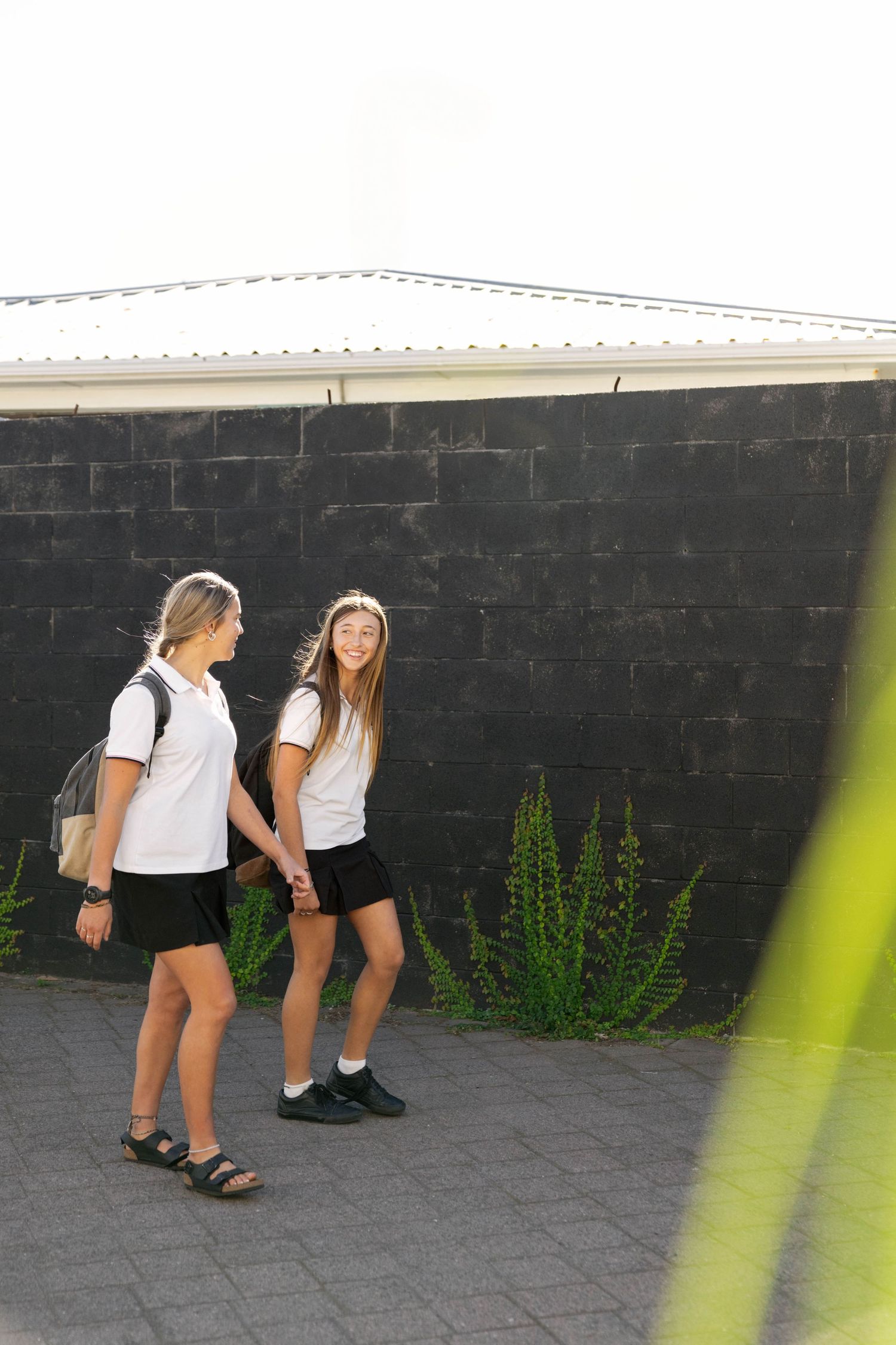 Two casual figures standing near a black wall on a paved outdoor area with plants growing nearby.