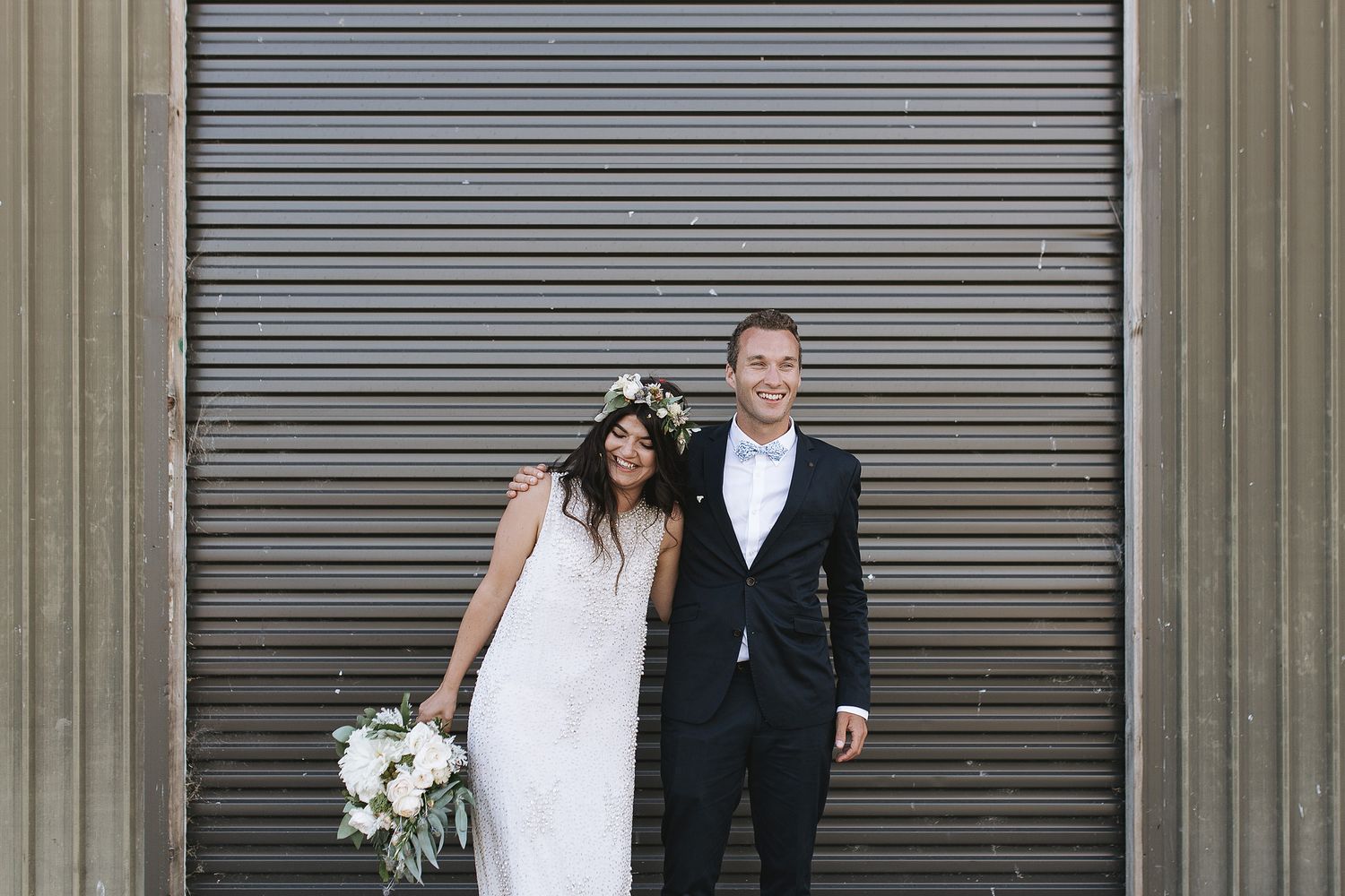 A couple walks down an urban street with white flowers and elegant attire in front of an industrial backdrop.