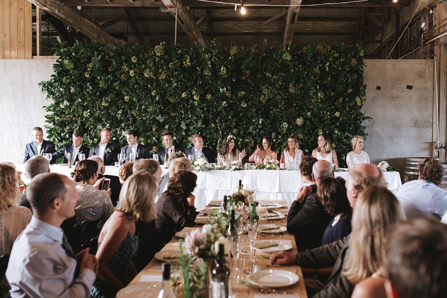 Wedding reception guests seated at long rustic wooden tables with greenery wall backdrop and elegant table settings.