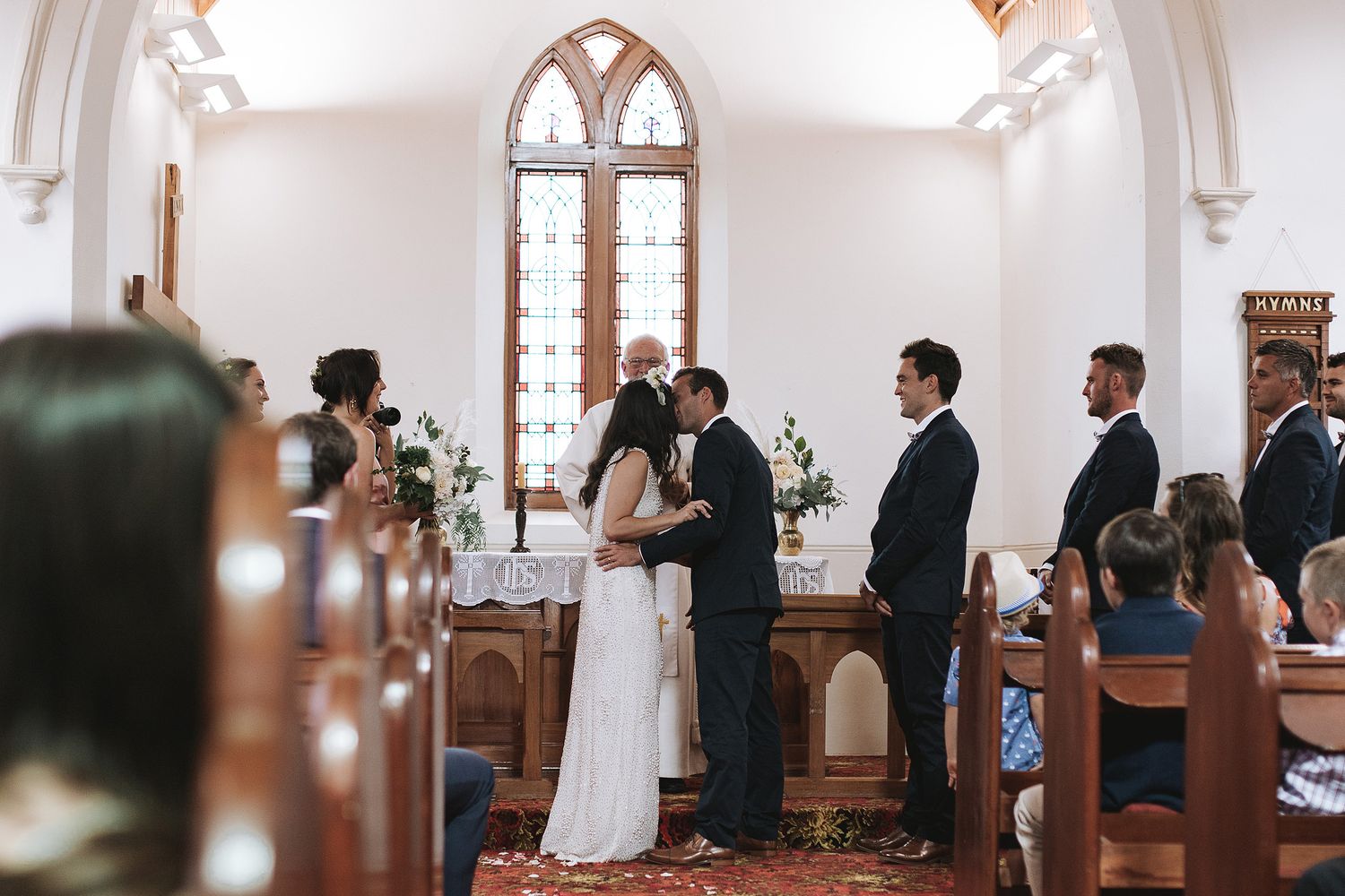 A bride and groom share their first kiss during a wedding ceremony in a classic white chapel with ornate arched windows.