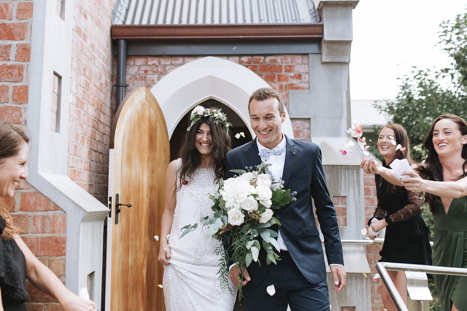 Happy newlyweds exit a brick church doorway as wedding guests celebrate with confetti in the background.
