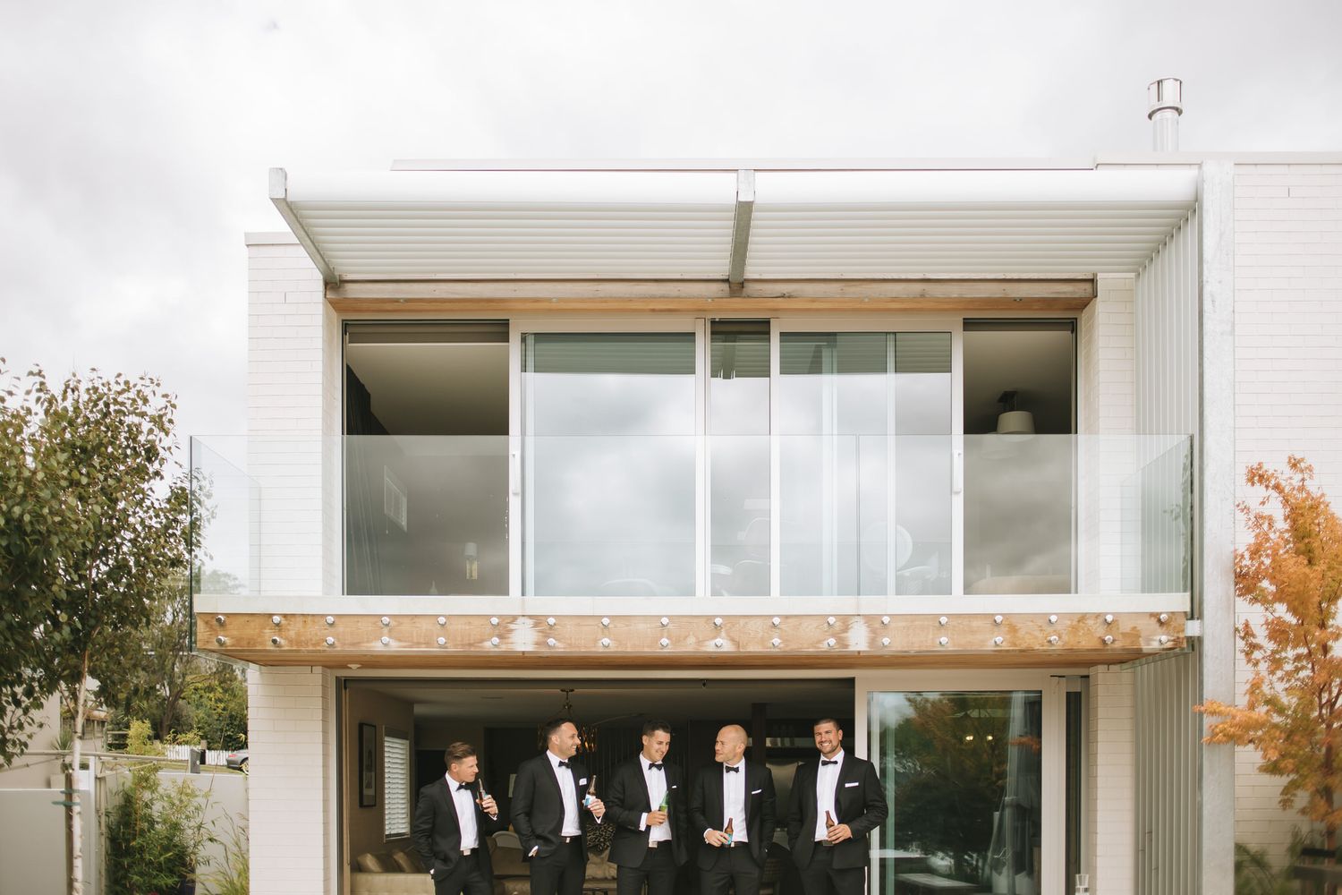 Wedding party dressed in dark suits stands in front of modern white building with floor-to-ceiling windows and autumn trees.