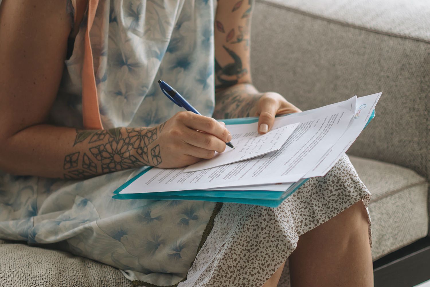 Person writing in a notebook while sitting on a grey couch with papers and documents spread around them.