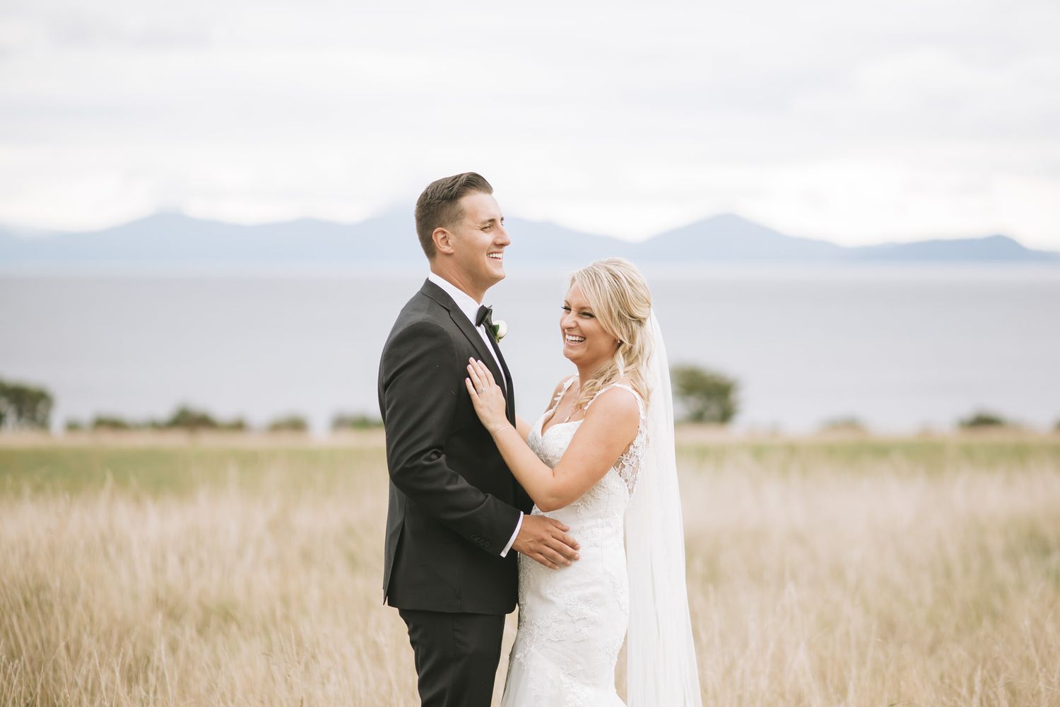 A couple shares a joyful moment in their wedding attire while standing in a golden field with mountains in the background.