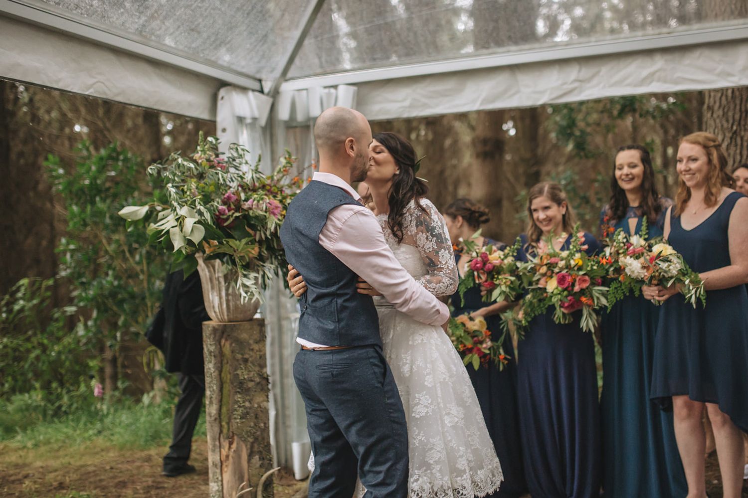 A romantic outdoor wedding ceremony under a canopy with bridesmaids in navy dresses holding colorful bouquets.
