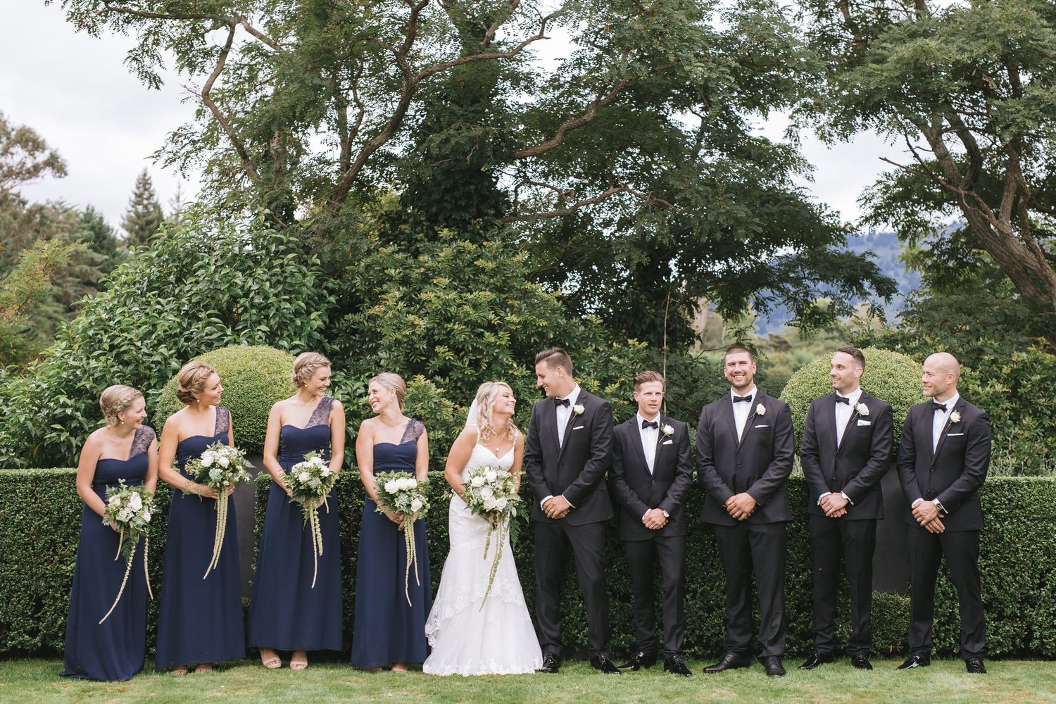 Wedding party in navy blue dresses and black tuxedos poses together on lawn with green hedge backdrop.