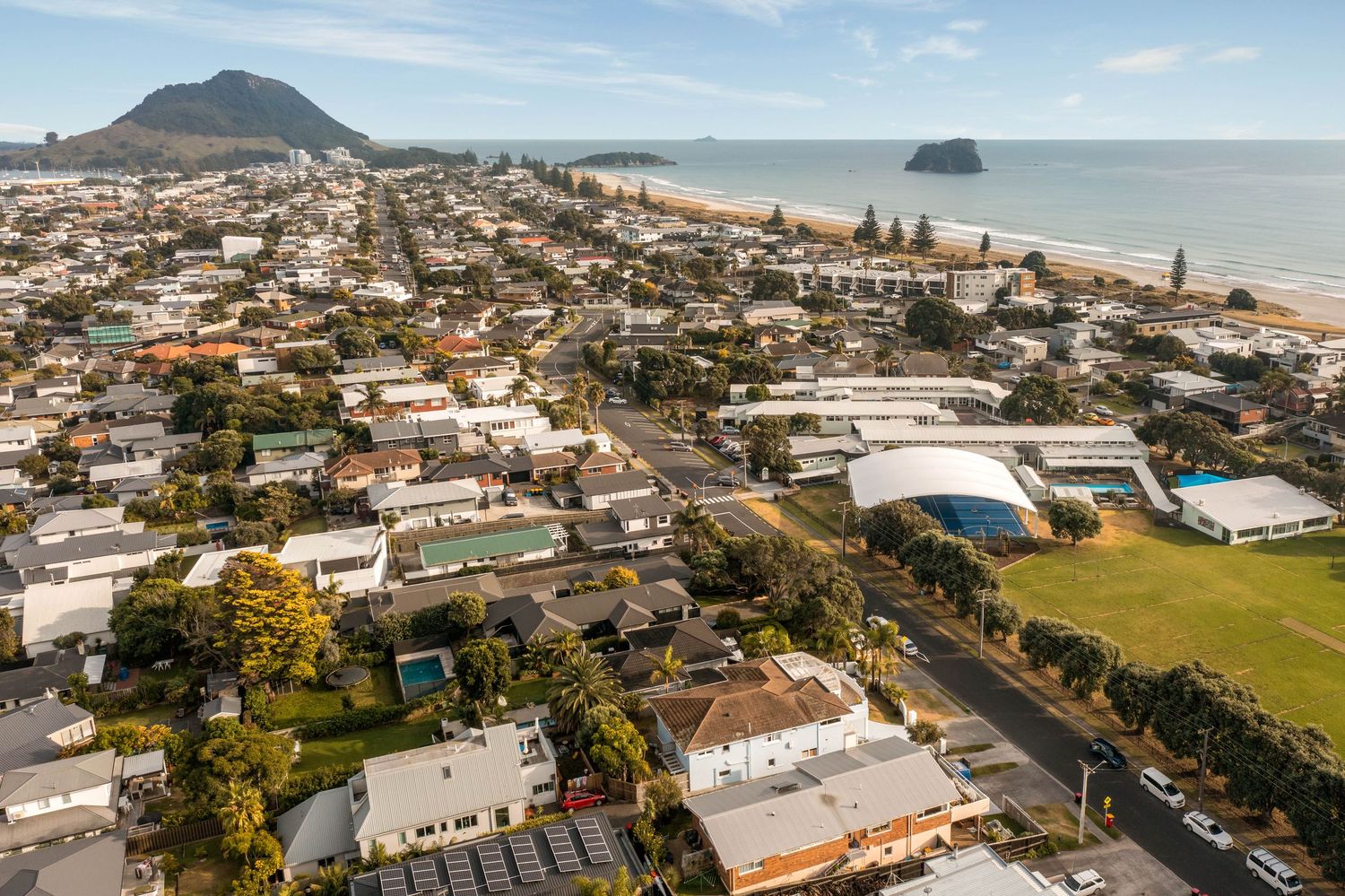 Aerial panoramic view of Mount Maunganui beach town with ocean, mountains, and coastal buildings stretching along shore.
