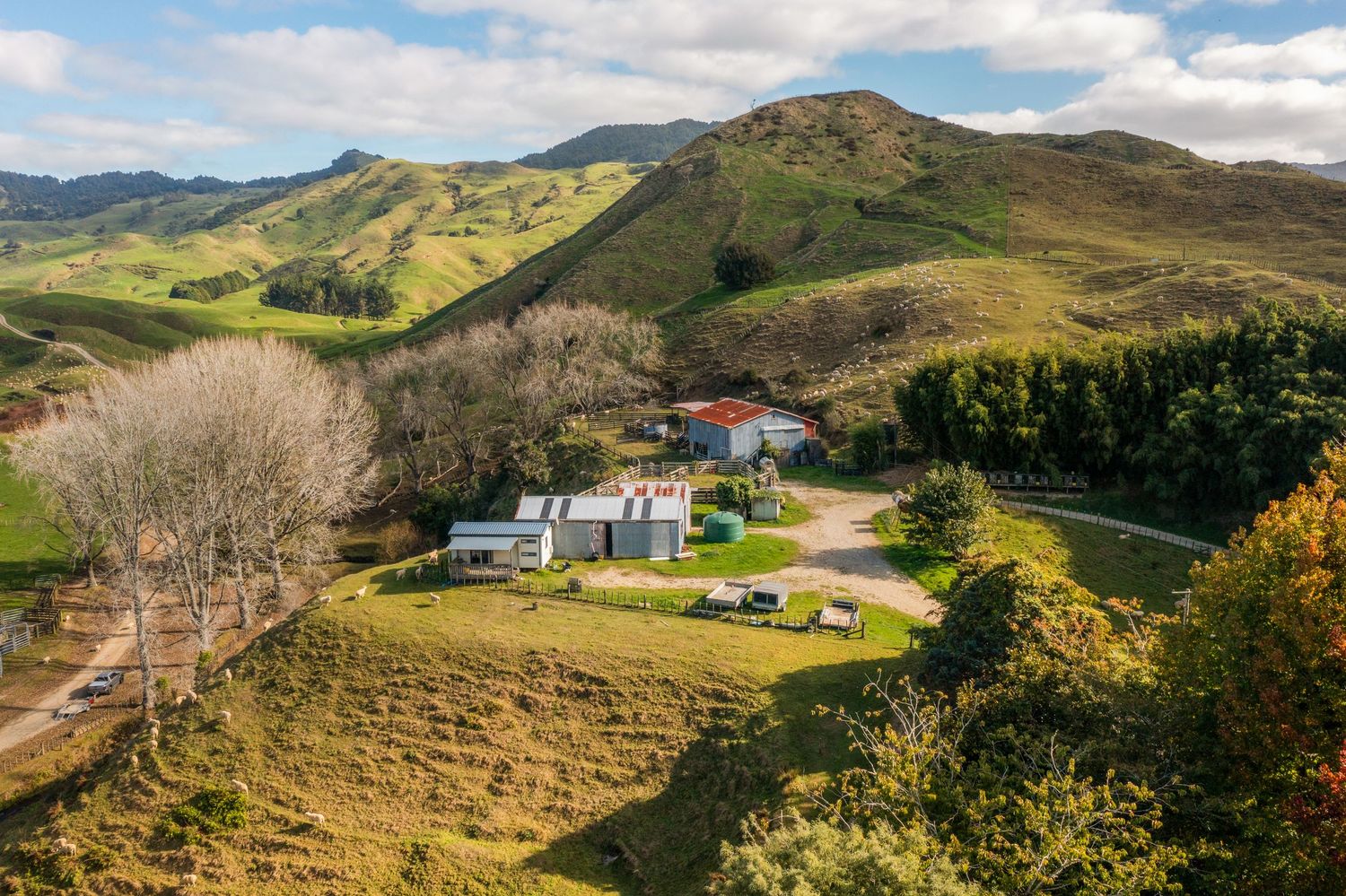 Rural property with buildings nestled in rolling green hills under cloudy blue sky.