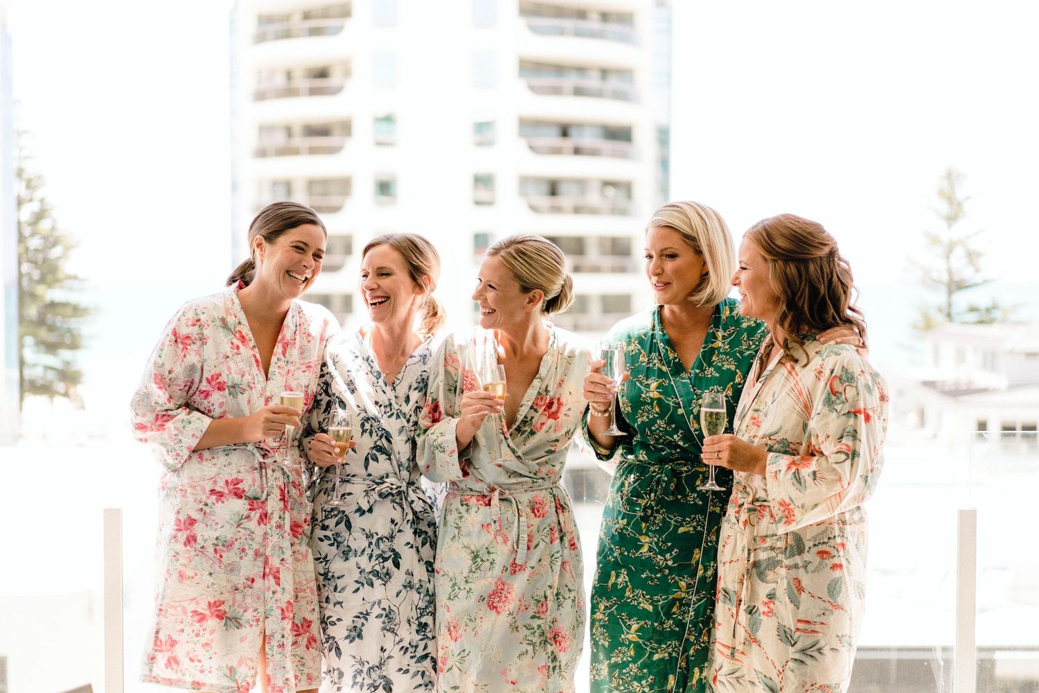 Five friends share a joyful moment together while wearing floral robes outdoors on a sunny day.