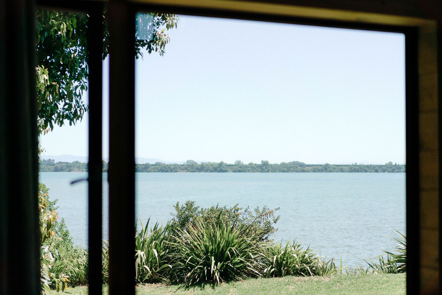 Scenic waterfront view through large windows with coastal vegetation in foreground.