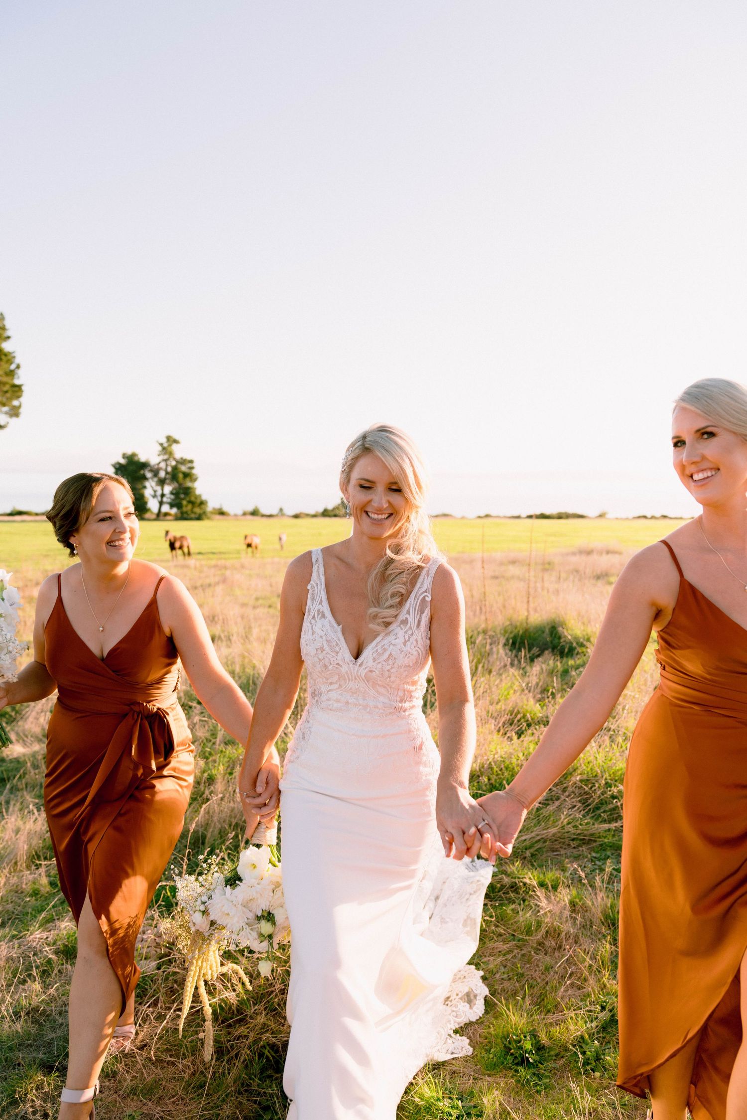 Three women in formal dresses walk together through a sunlit field at sunset.