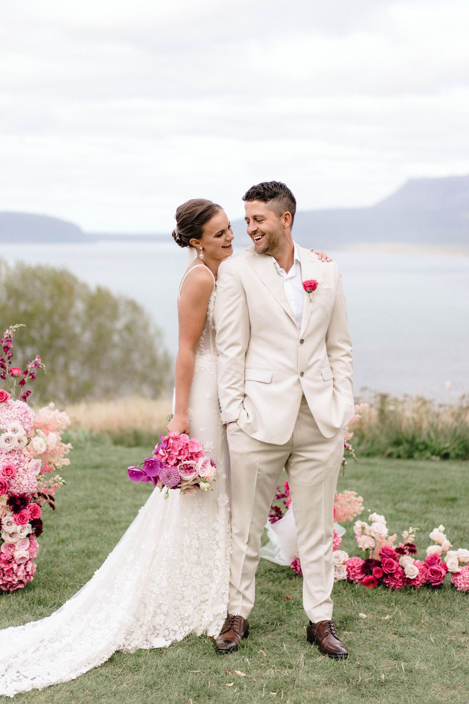 Happy couple shares romantic moments in their beige wedding attire surrounded by pink flowers near an oceanside meadow.
