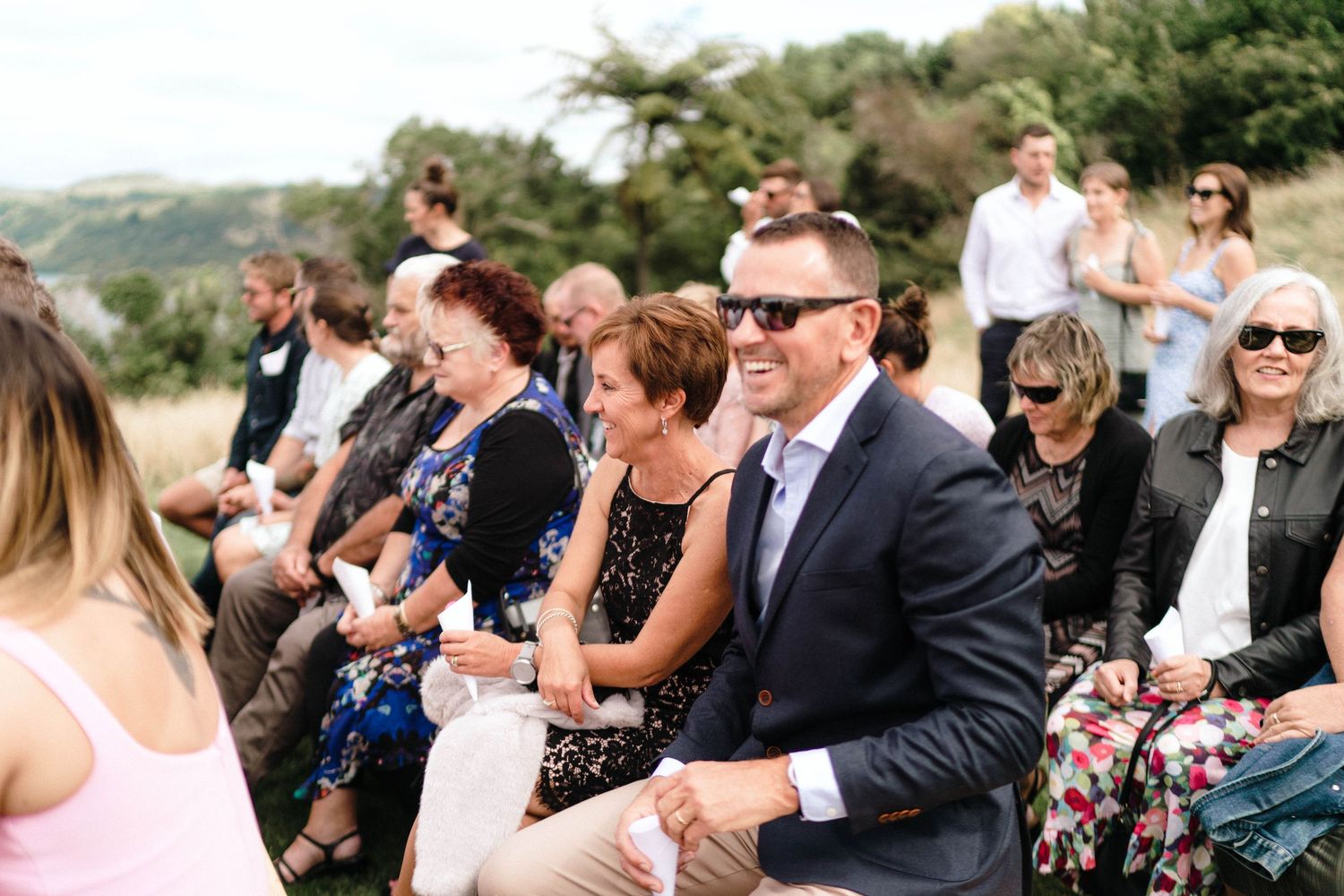 Wedding guests in formal attire seated outdoors watching a ceremony on a sunny day.
