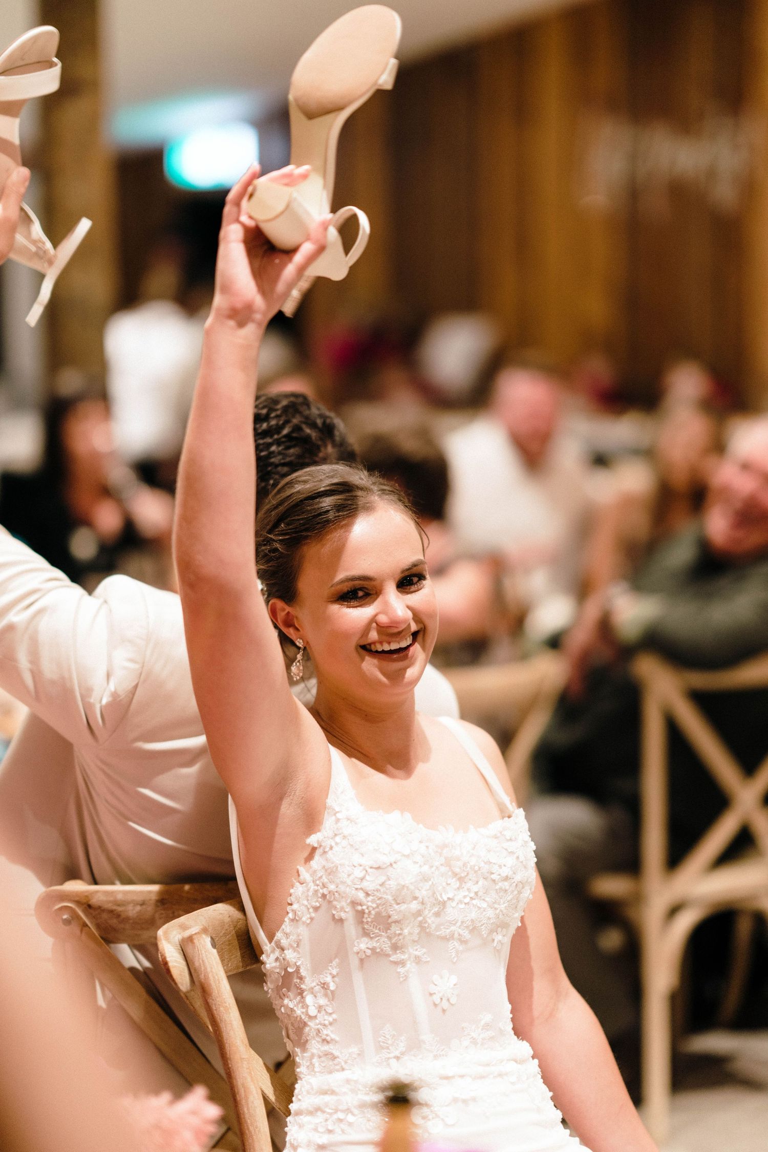A bride in a white wedding dress celebrates joyfully with raised arms during a reception celebration.