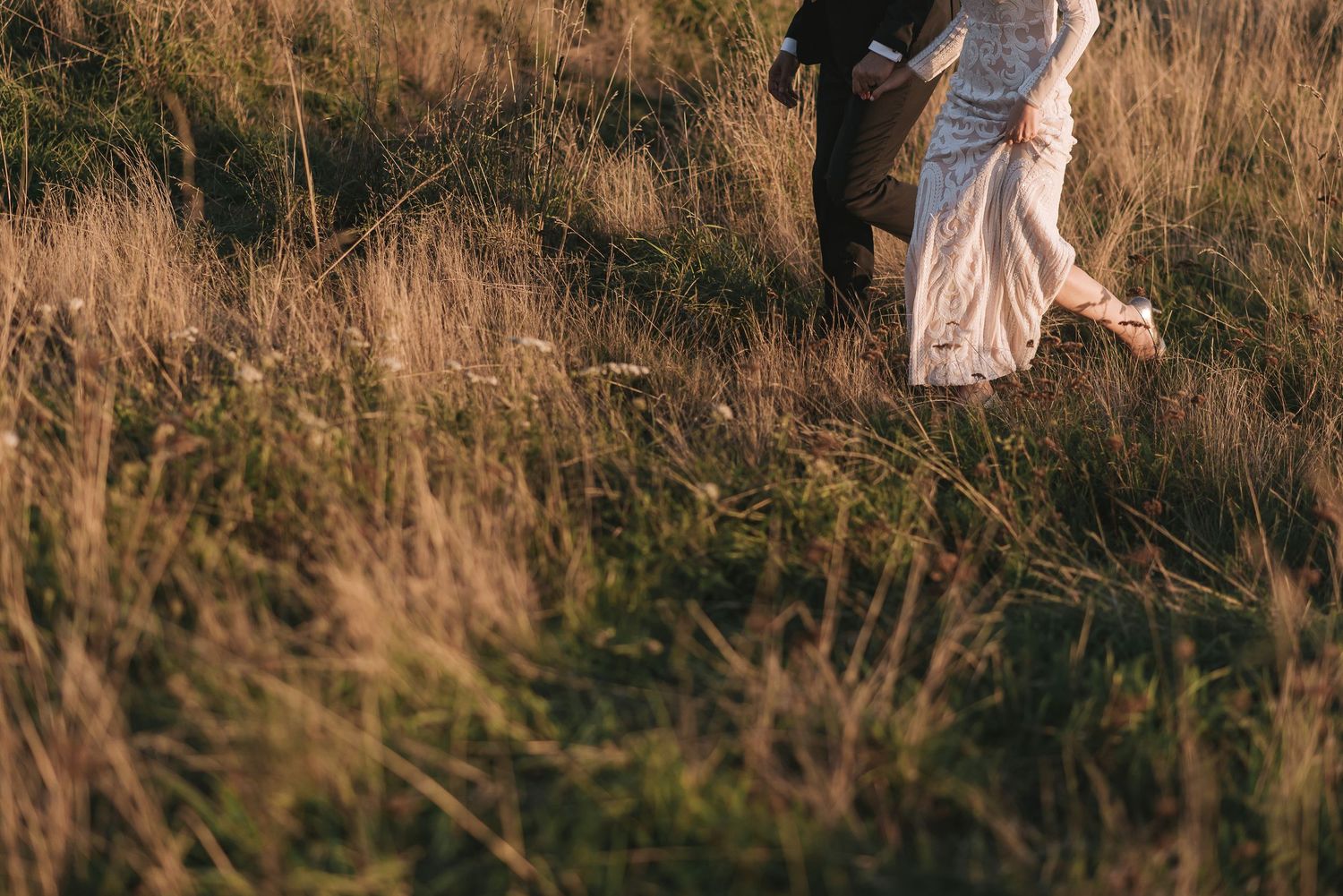 A couple holds hands while walking through a sunlit field of tall golden grass during sunset.