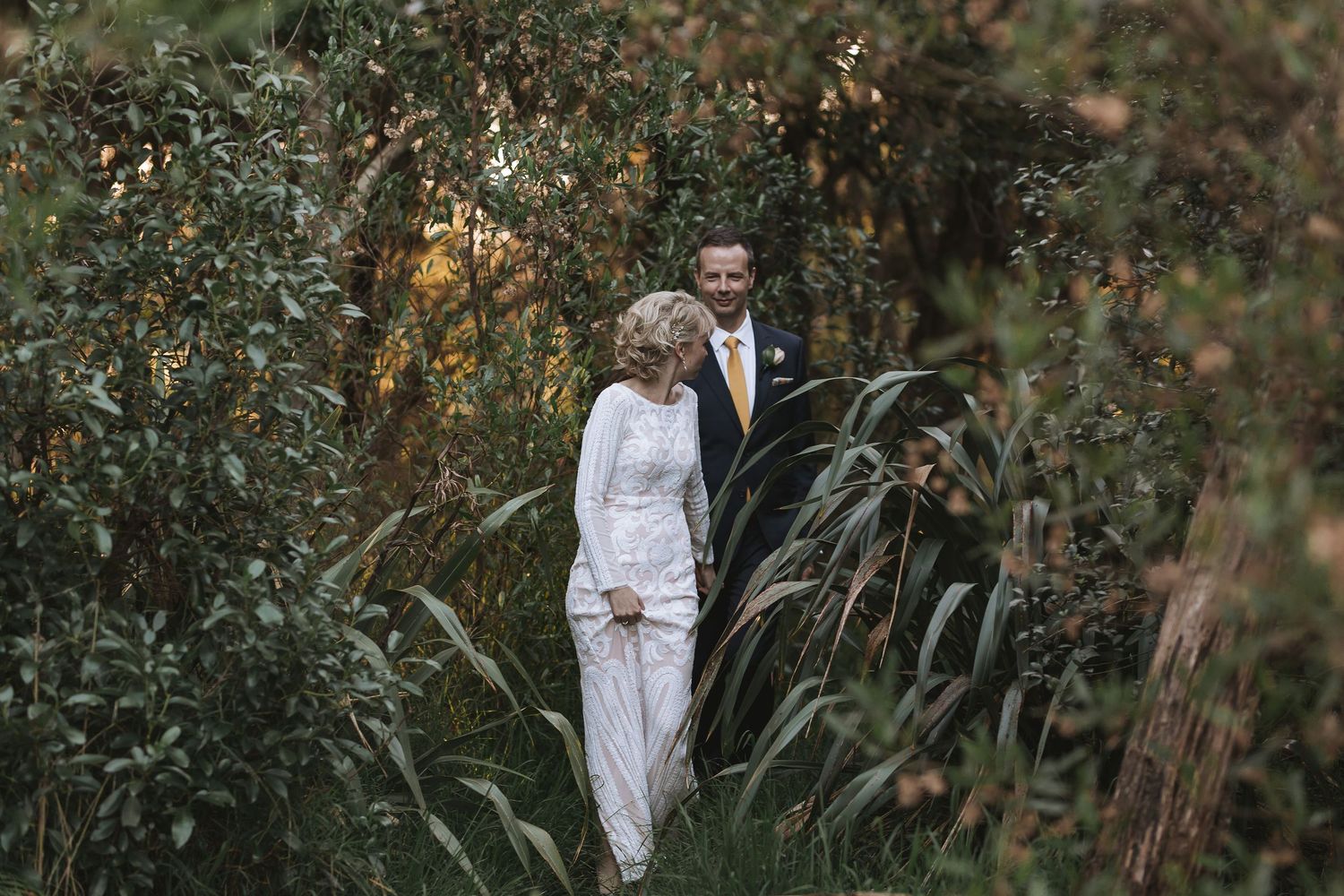 A bride and groom share a tender moment in a lush garden setting with natural backlit sunlight filtering through trees.