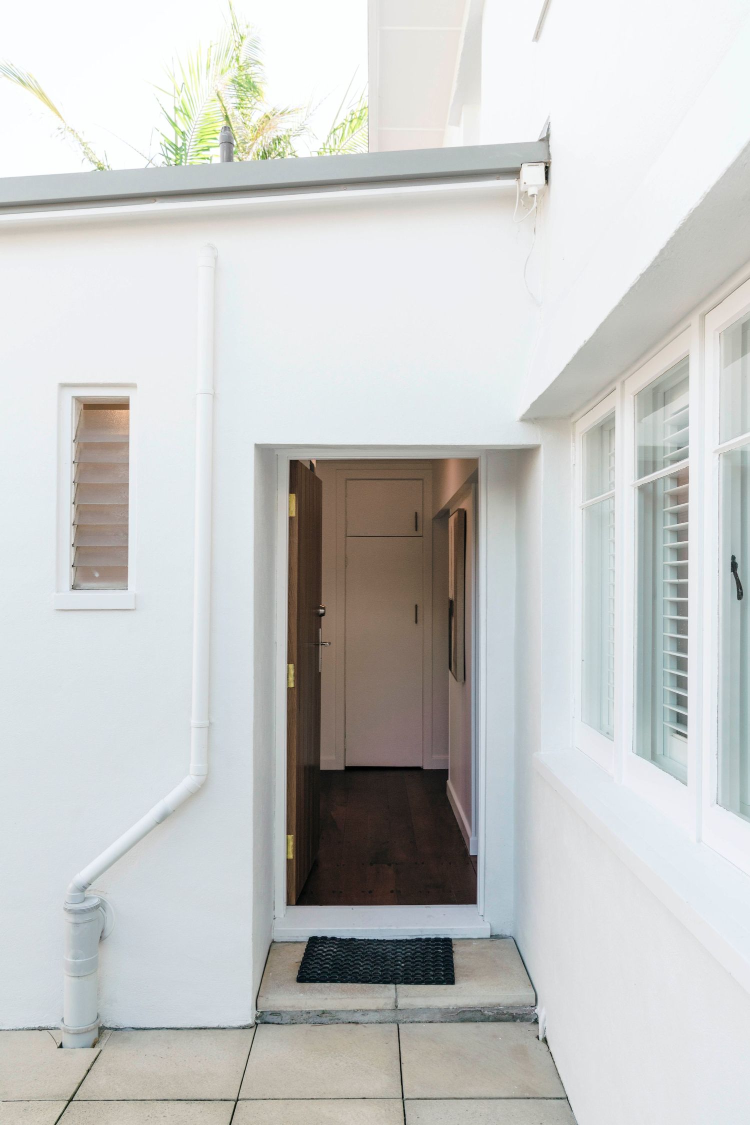 White exterior entrance with shuttered windows, tiled floor and dark doormat leading to a hallway.