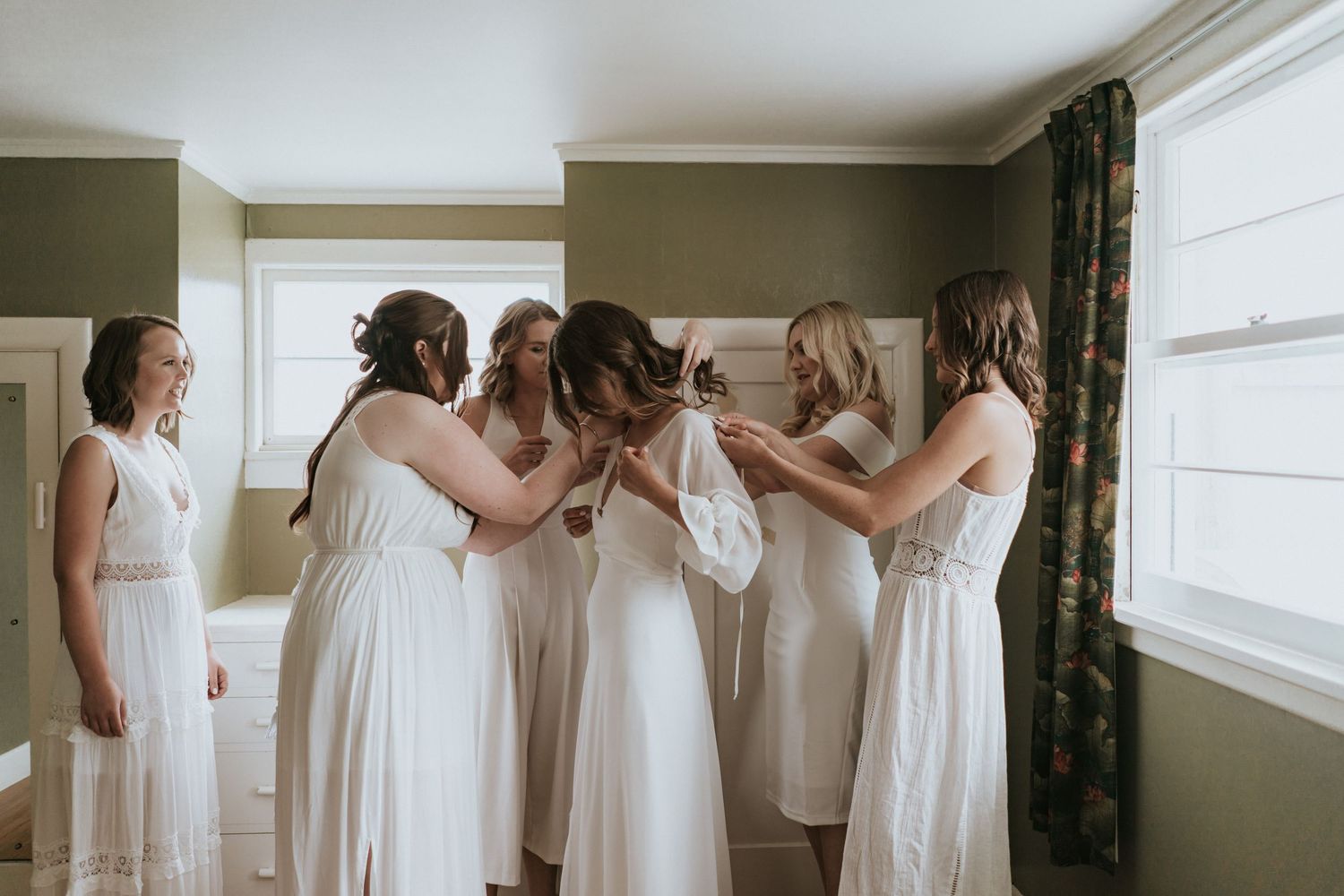 A group of bridesmaids in white dresses help each other get ready in a sunlit room before the wedding ceremony.