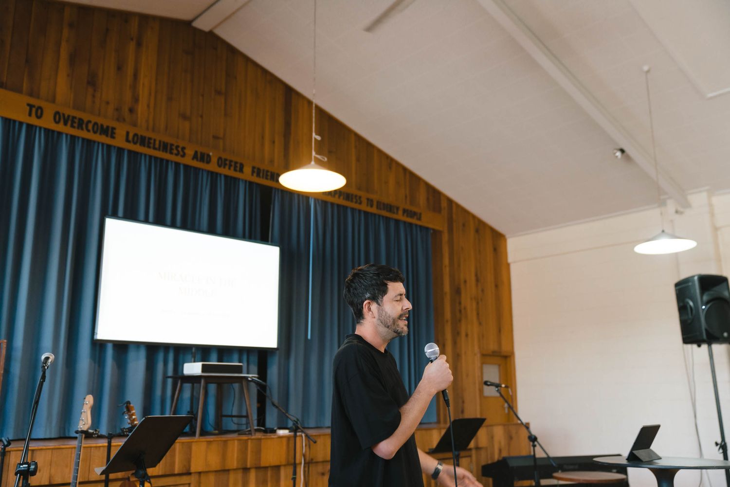 Speaker presenting on stage with projection screen and audio equipment in church-style auditorium.