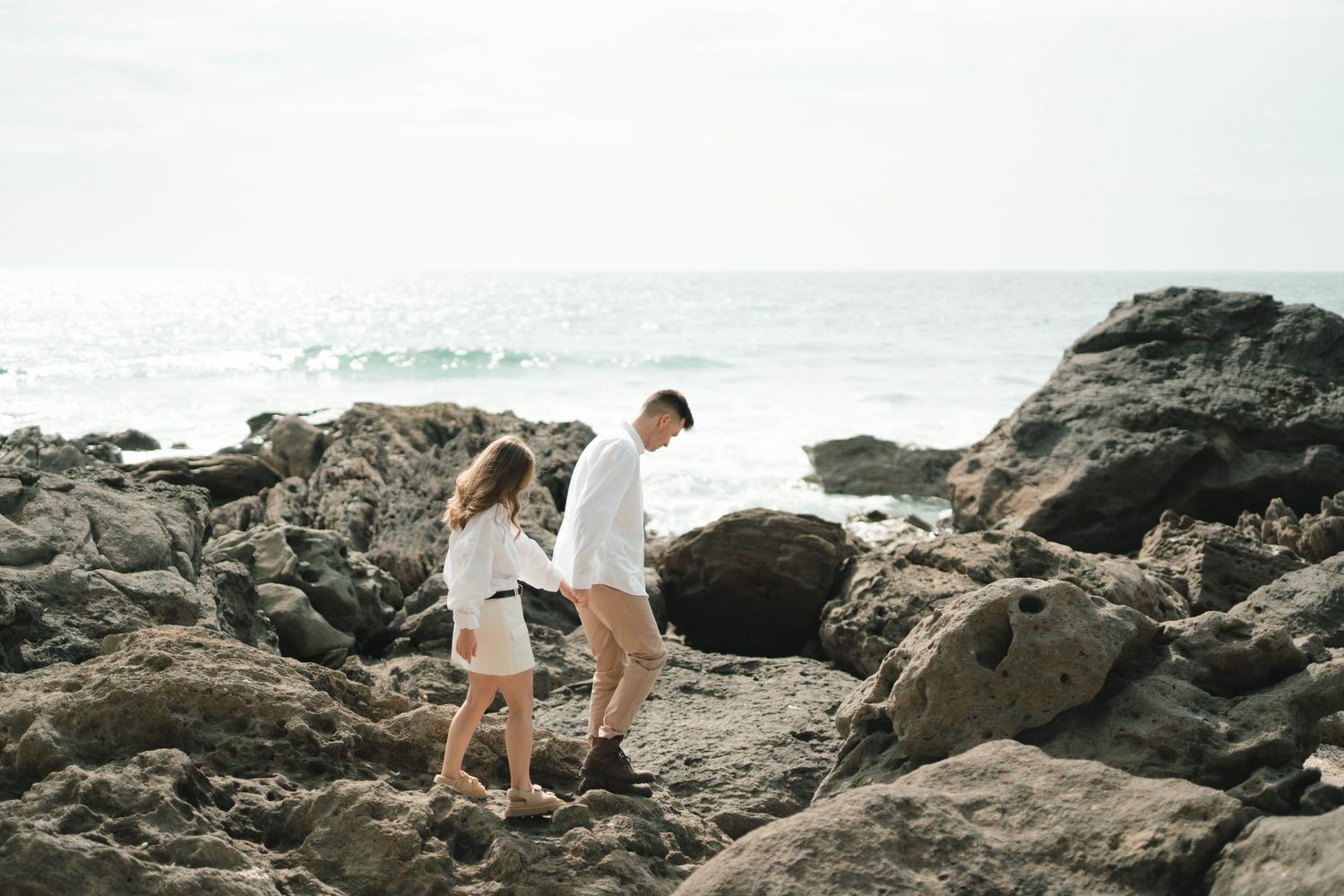 Two people carefully explore rocky coastal terrain against a misty ocean backdrop on a cloudy day.