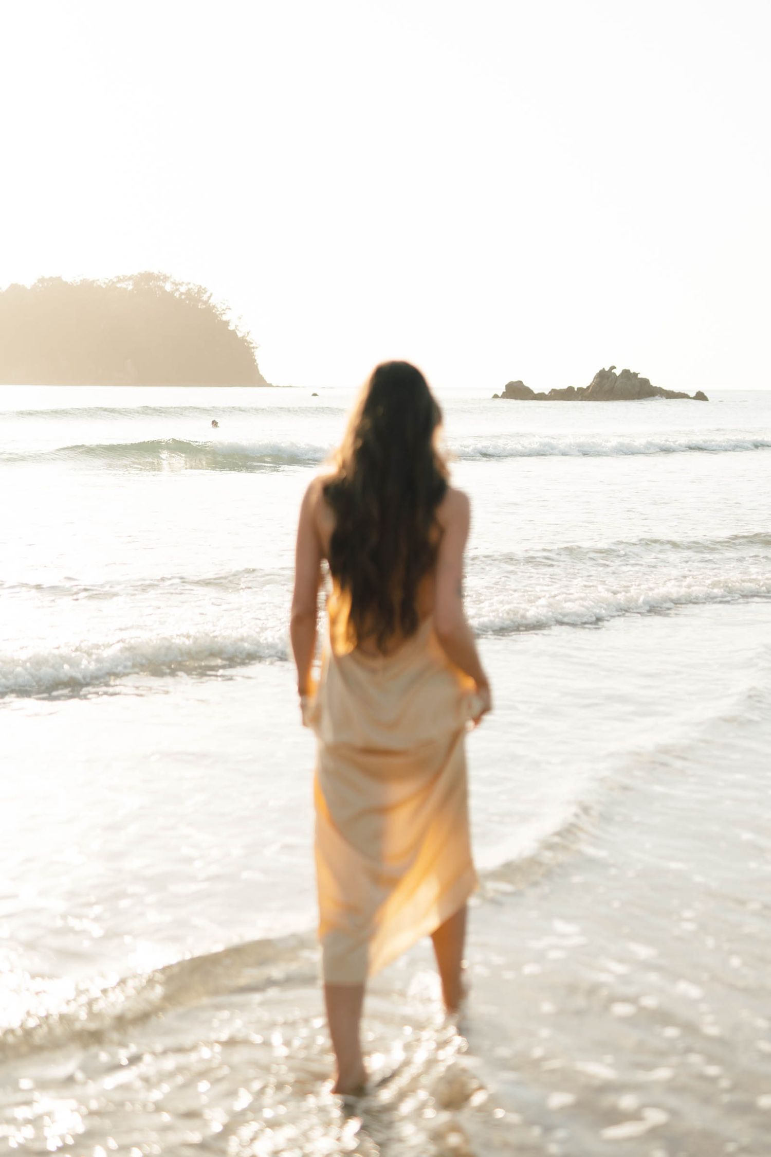 Figure in flowy white dress walks along ocean shoreline at sunset.