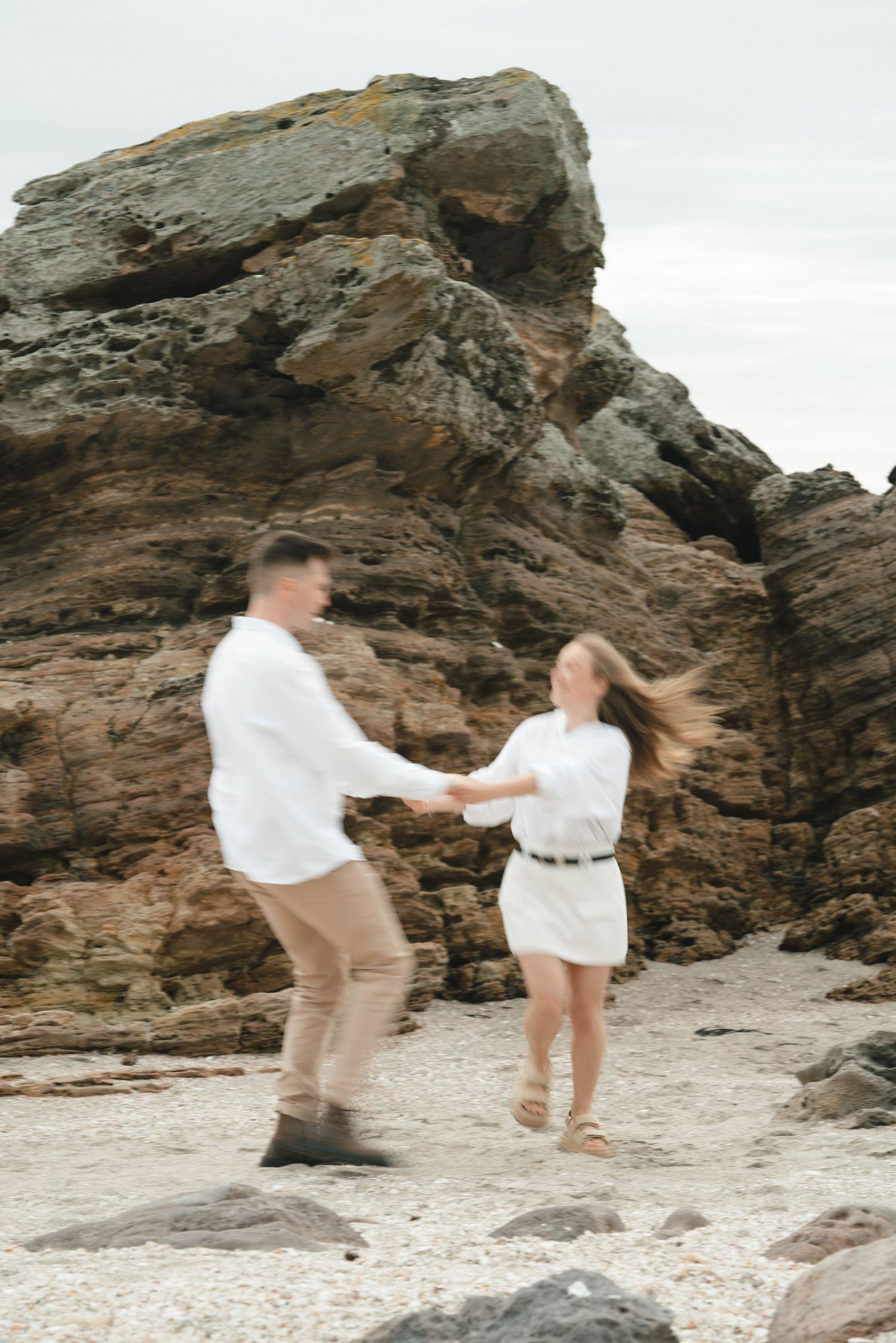 Couple in white clothing dancing together on a rocky beach.