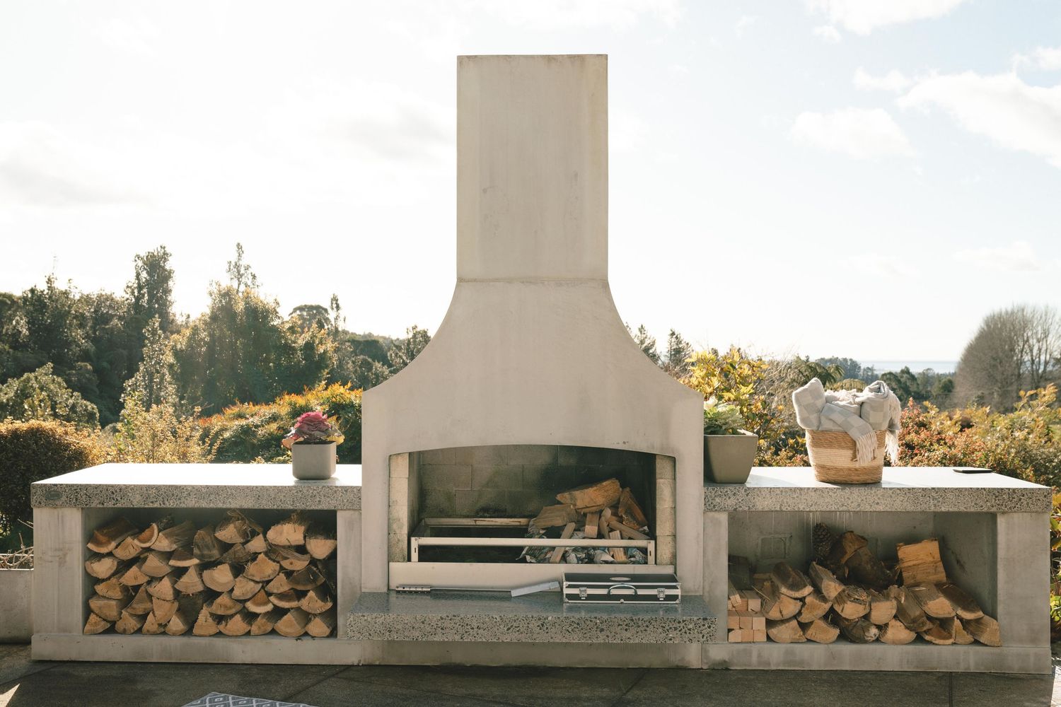 Modern outdoor concrete fireplace with built-in firewood storage and seating area surrounded by lush garden setting.