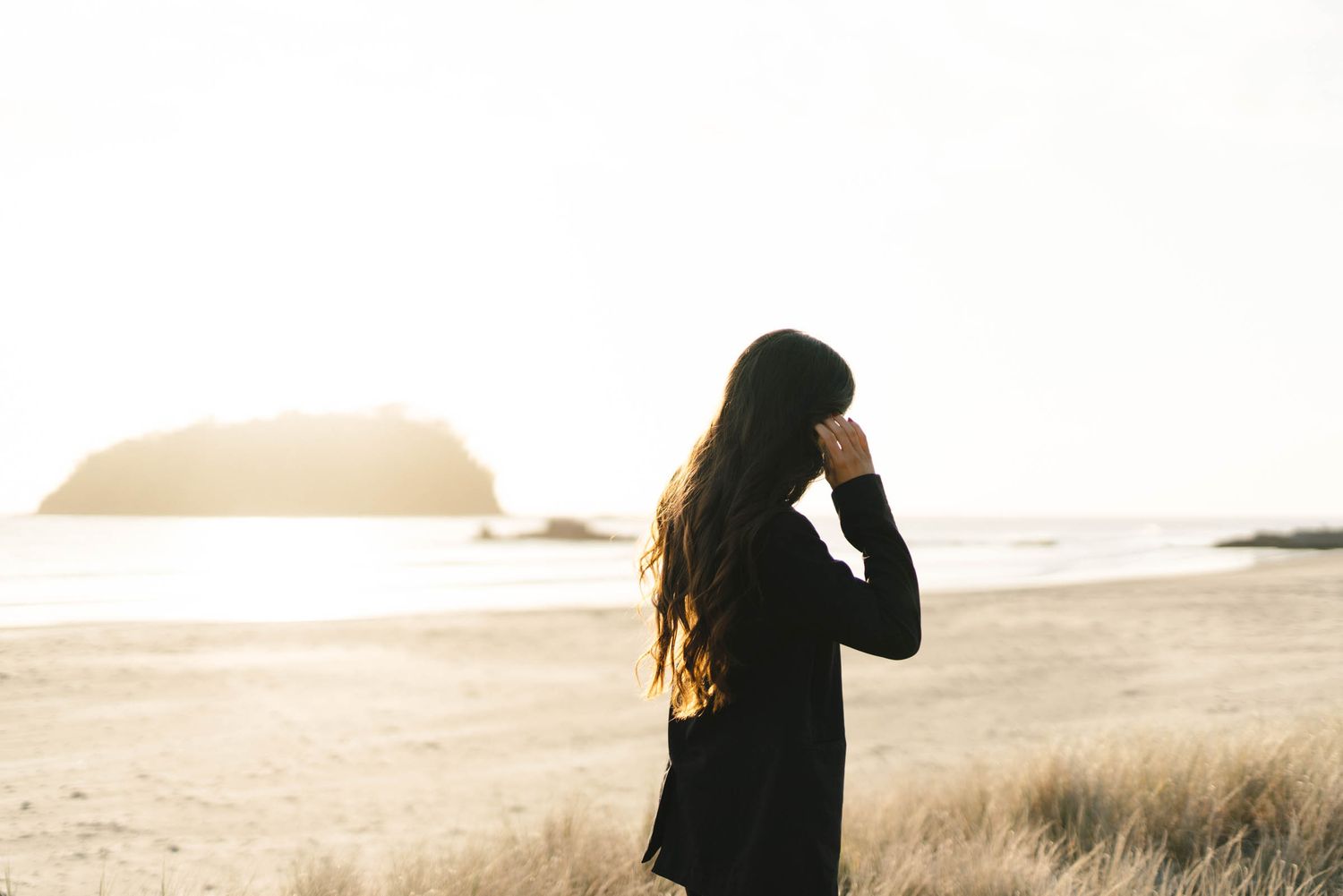 Silhouette of a woman in black clothing standing on a beach at sunset with windswept hair.