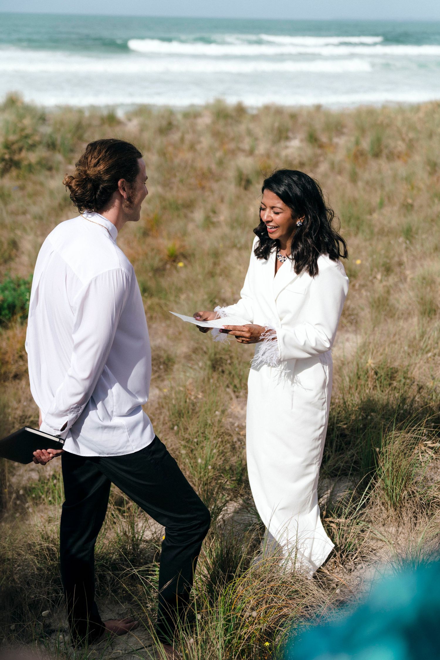 A couple playfully interacts on a grassy beach hillside wearing white and black outfits.