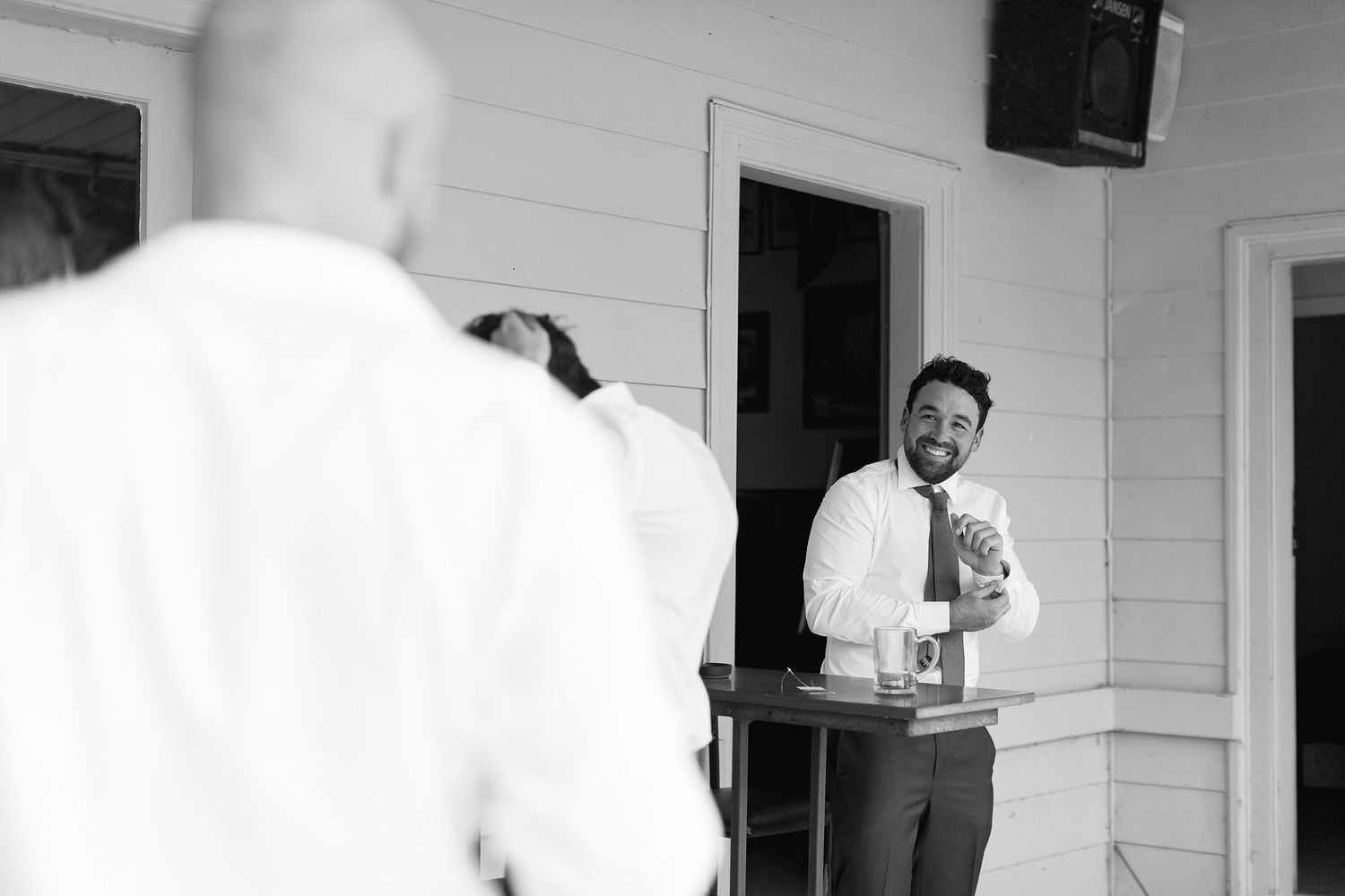 Black and white photo of someone in a dress shirt and tie standing in a doorway of a white house, gesturing with their hand.