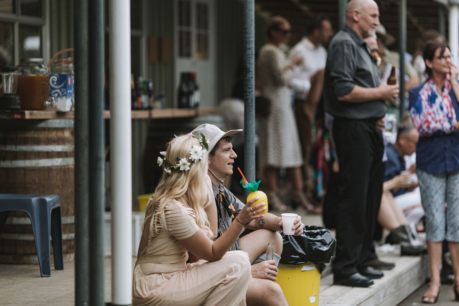 Two festival attendees in summer attire sit and enjoy drinks on steps while others stand in background.