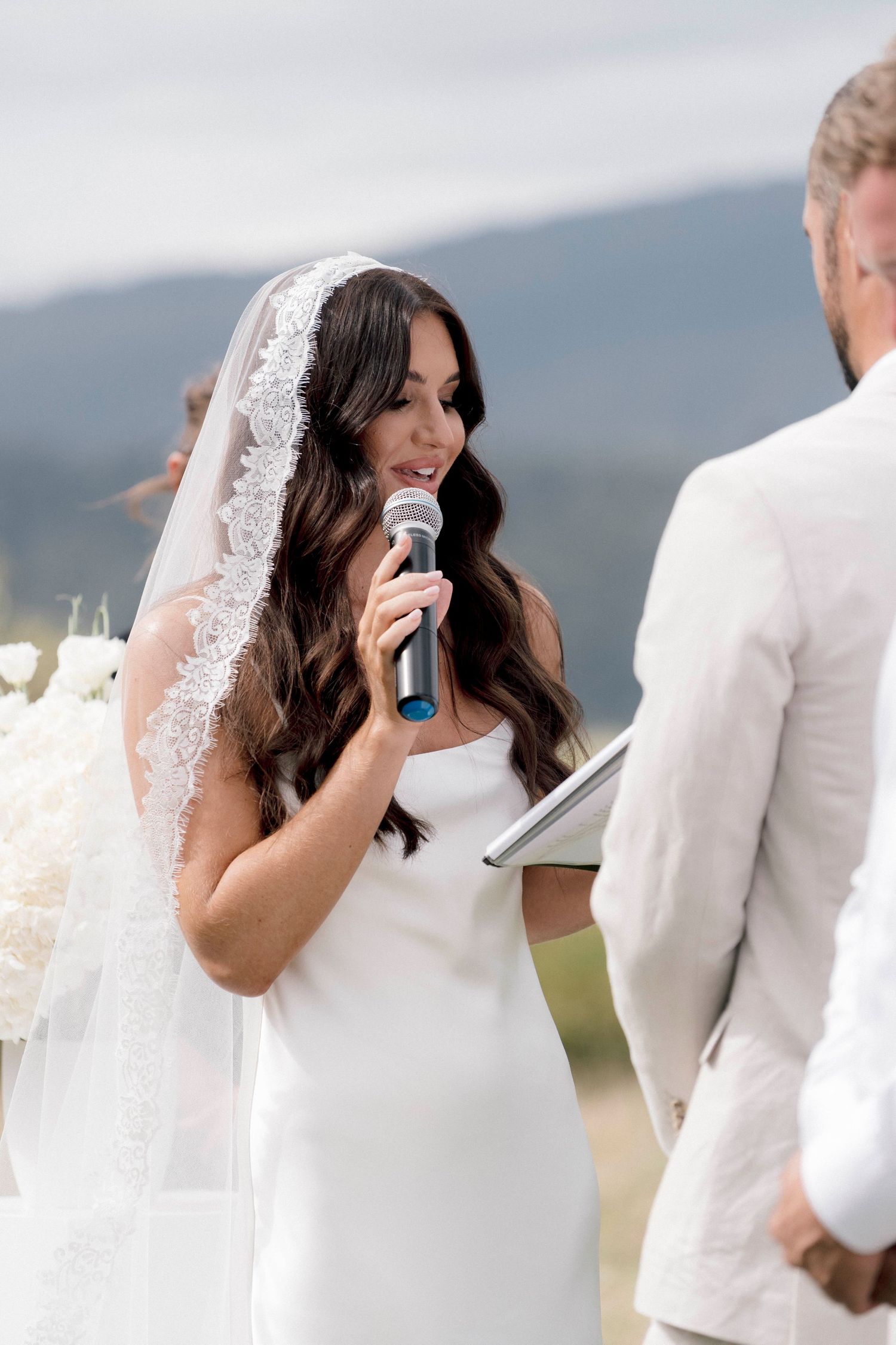 Bride in a white wedding dress and veil reads vows from paper while holding microphone during outdoor mountain ceremony.