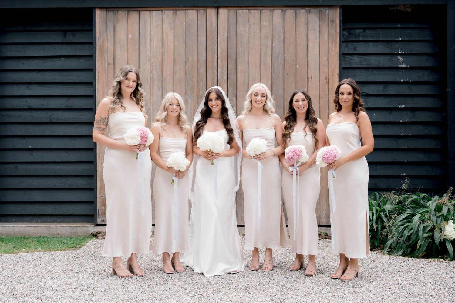A bridal party in cream-colored dresses poses in front of a dark barn building.