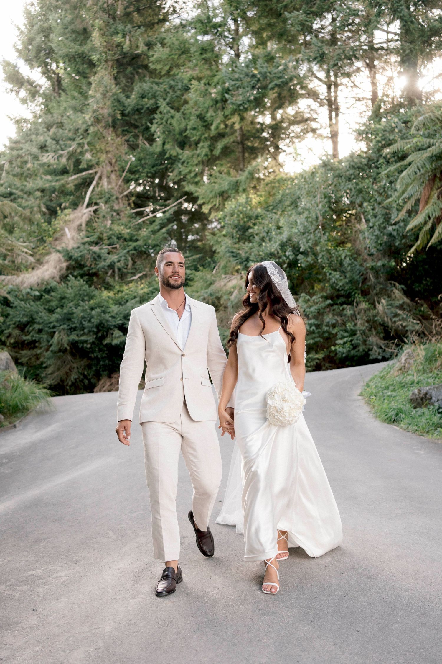 A newlywed couple walks down a tree-lined road wearing elegant all-white wedding attire.