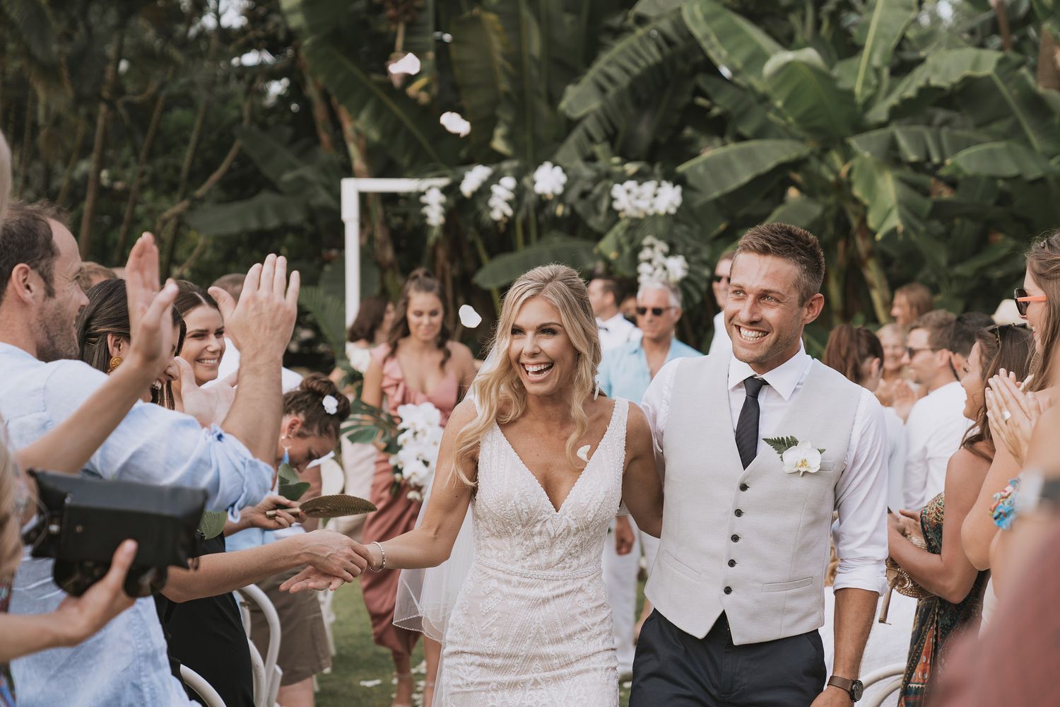 Joyful wedding guests dance and celebrate during an outdoor reception with tropical greenery backdrop.