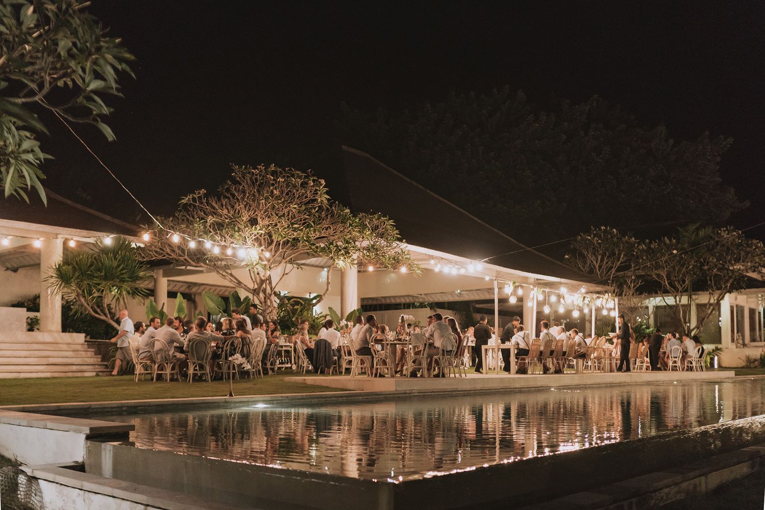 Elegant outdoor wedding reception by a reflective pool with string lights and guests dining under a white tent.
