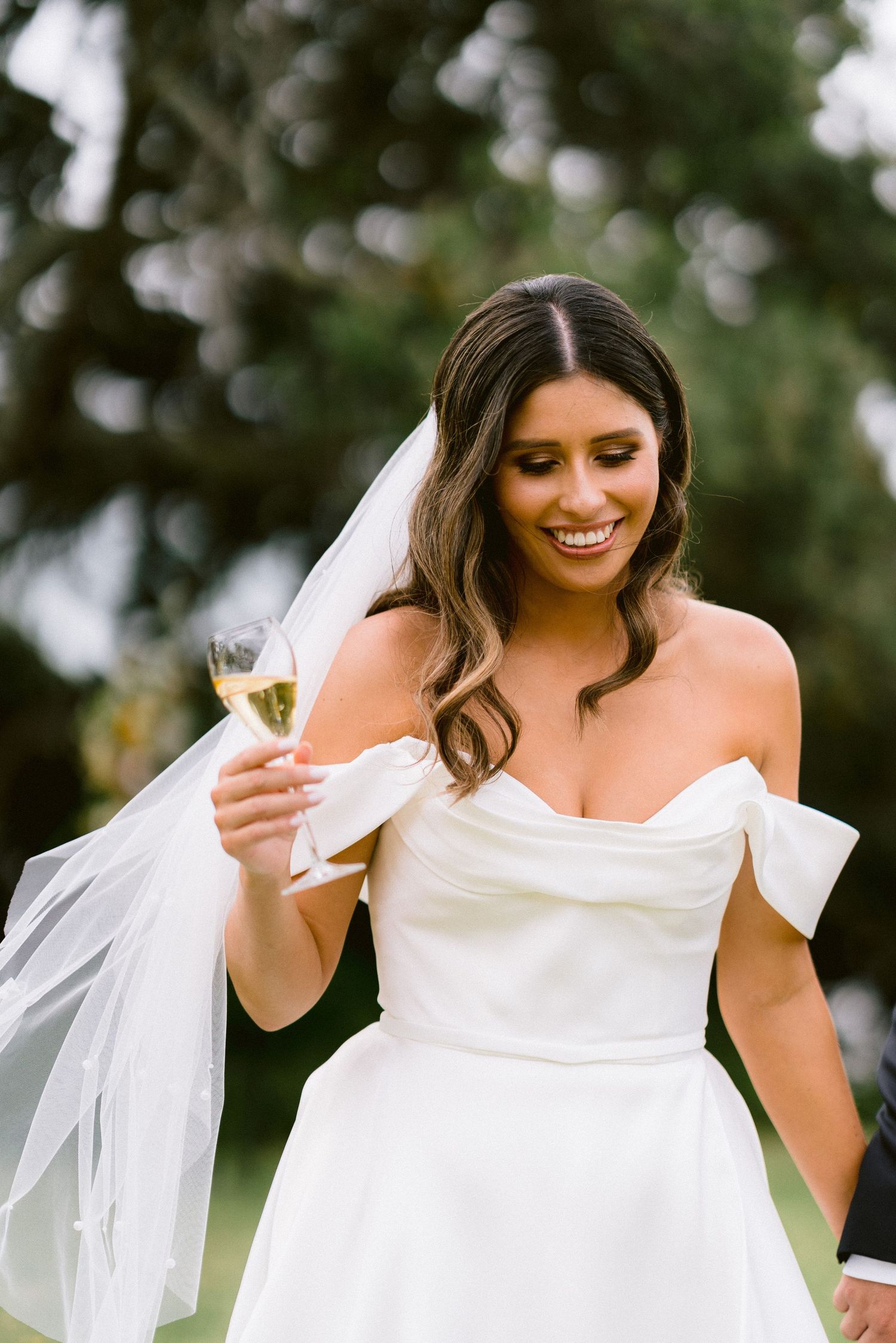 A bride in an off-shoulder wedding dress holds a champagne glass while smiling.