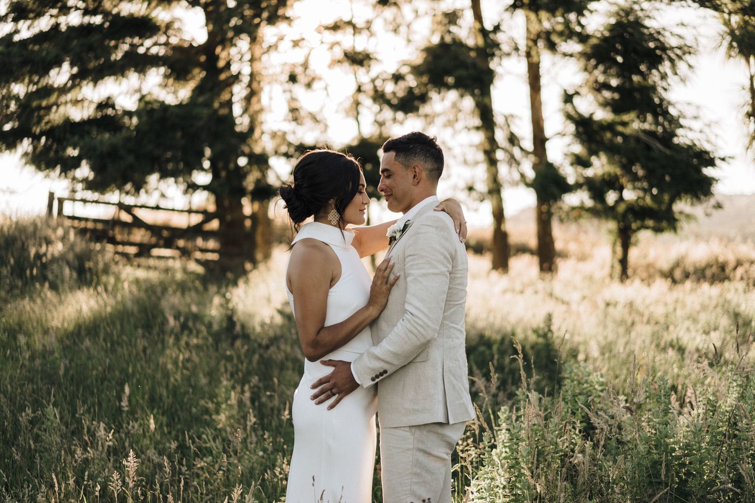 Couple in wedding attire embracing in golden sunset light with trees in background.