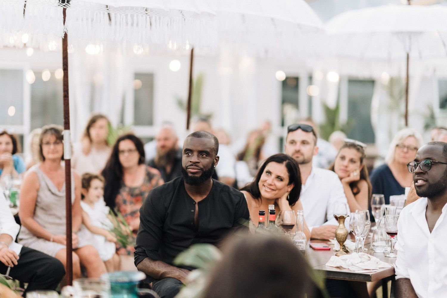 Diverse group of wedding guests gathered at outdoor reception tables beneath white tent decor.
