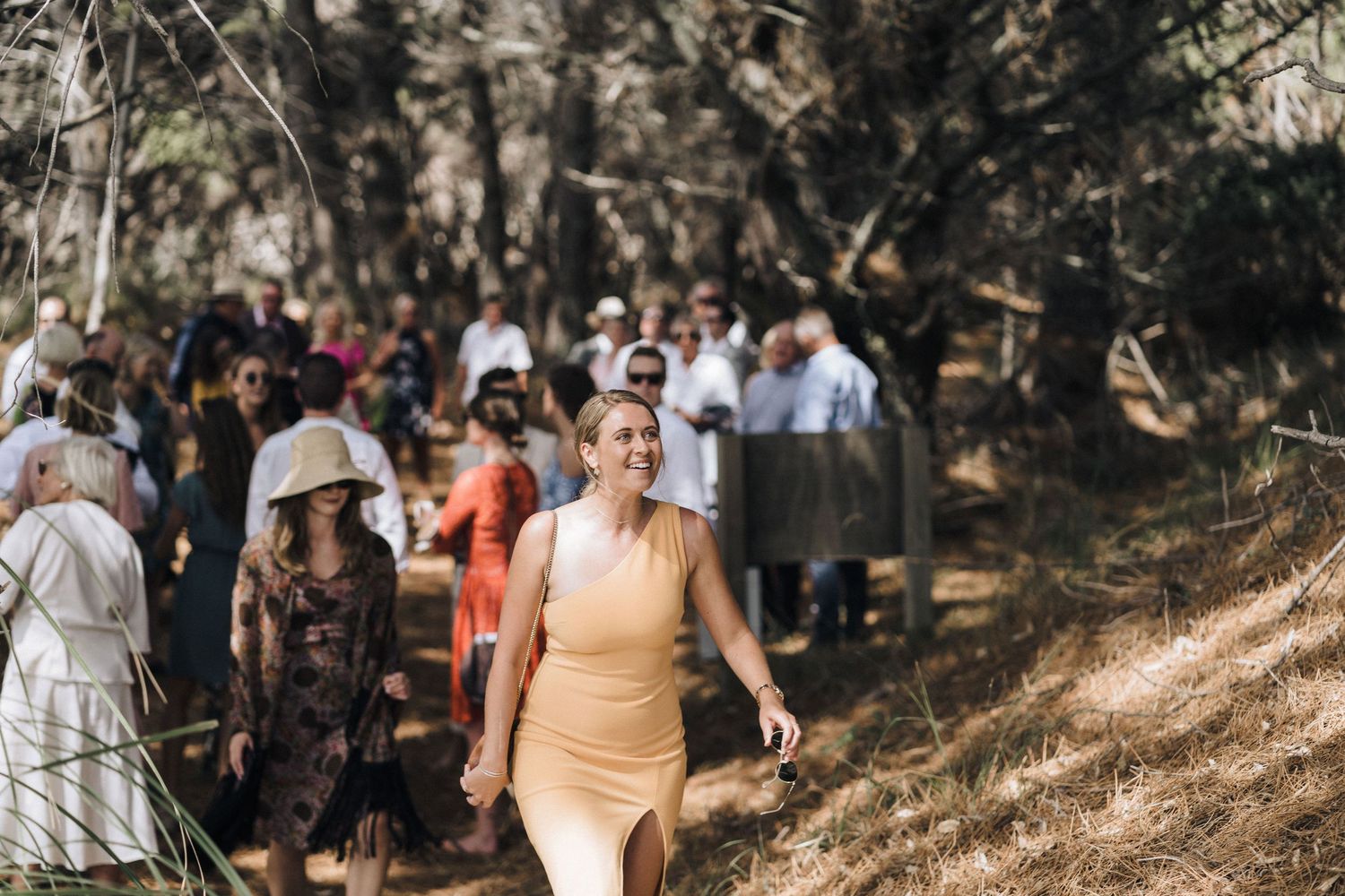 Two people in wedding attire walk through a wooded area surrounded by guests during an outdoor ceremony.