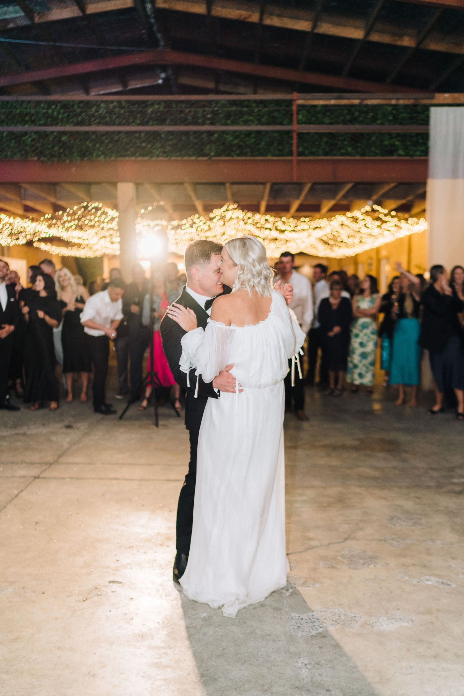 A first dance at a wedding reception under twinkling string lights with guests watching in the background.