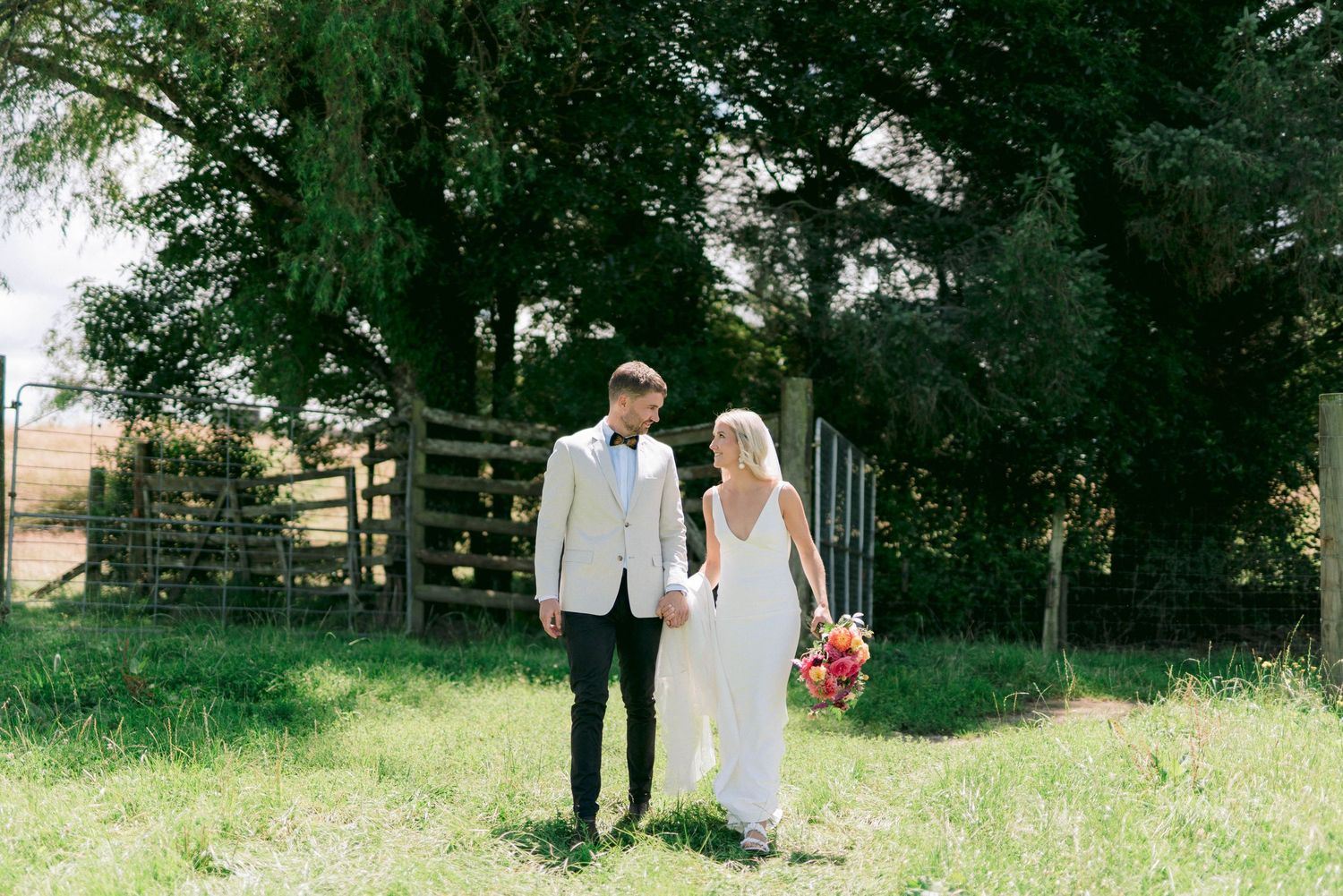 Bride and groom stand together on grassy path with rustic fence background.