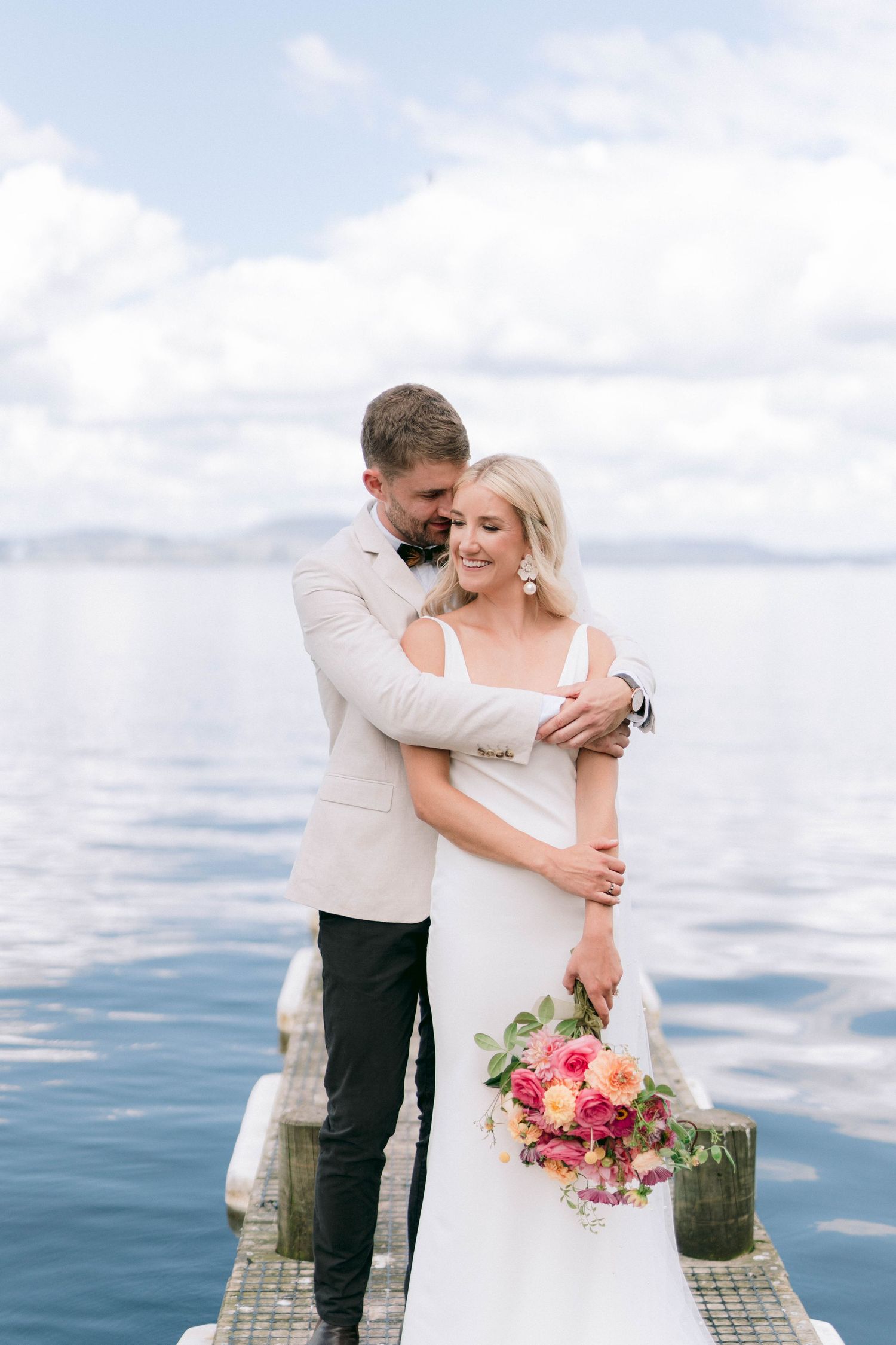 A romantic moment captured on a lakeside dock as newlyweds embrace with a vibrant bouquet.
