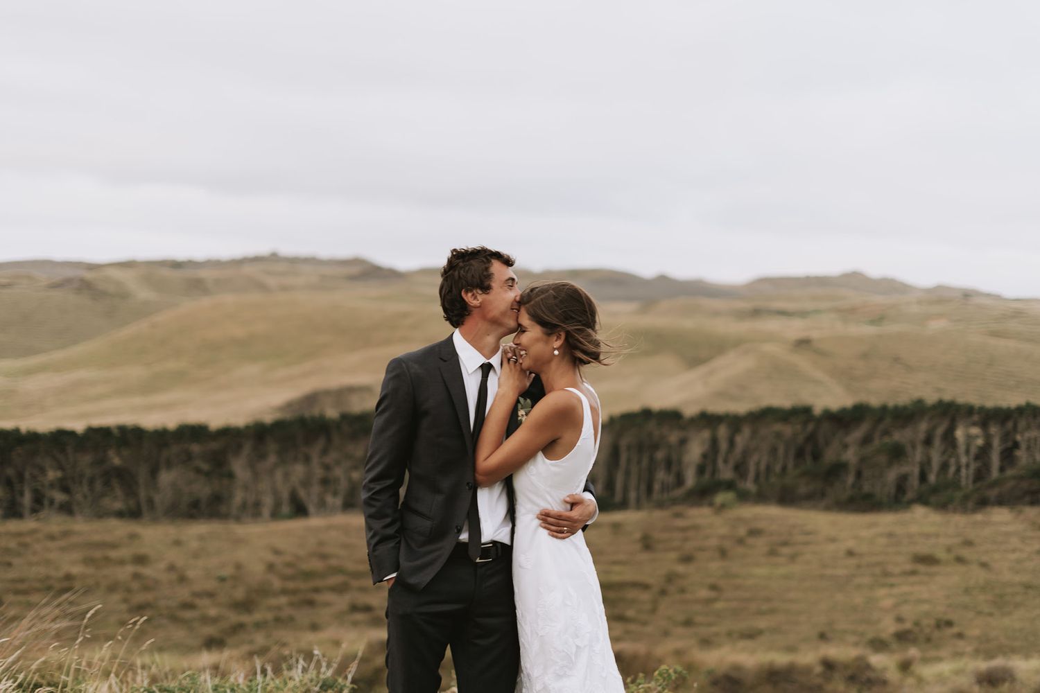 A couple embraces in their wedding attire against a scenic backdrop of rolling hills and cliffs at sunset.