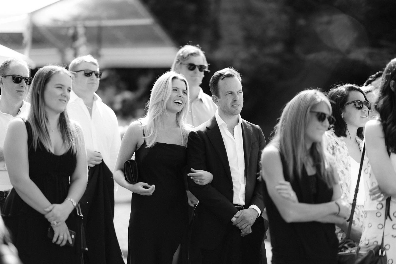 Black and white photo of outdoor wedding guests in formal attire gathered together watching a ceremony.