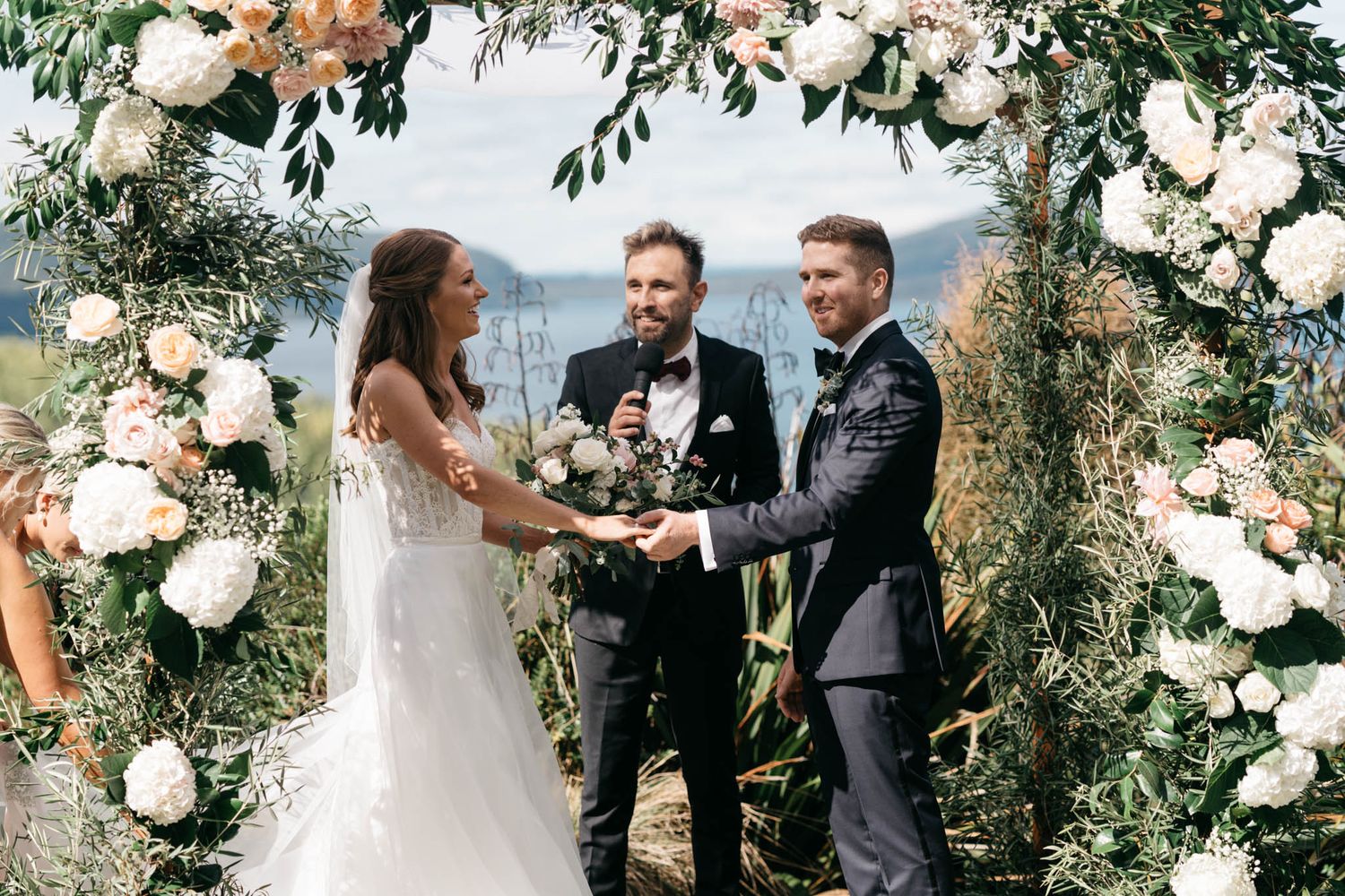 Wedding ceremony taking place under a beautiful floral arch with white roses and greenery against a scenic mountain backdrop.